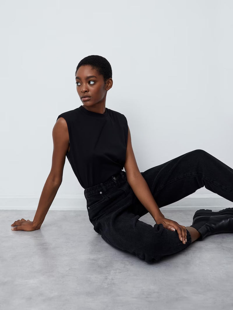Eine Person mit kurzen Haaren sitzt auf dem Boden vor einer weißen Wand. Sie trägt das Top „Kim“ von LeGer by Lena Gercke, kombiniert mit schwarzen Jeans und Stiefeln. In dieser minimalistischen Umgebung blickt sie nachdenklich zur Seite.