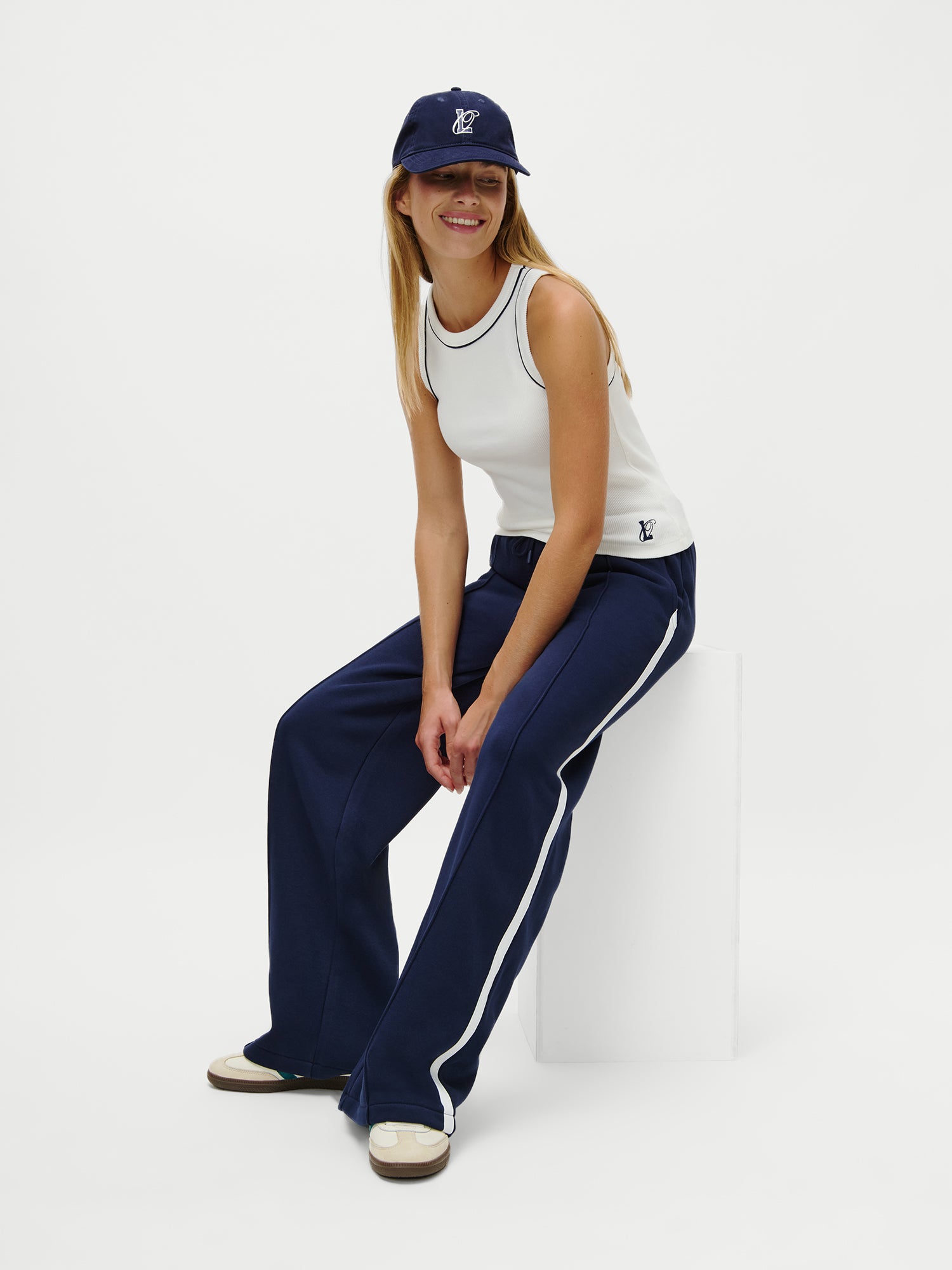 Eine Frau trägt das LeGer x Oh April - Dira Top von LeGer by Lena Gercke - ein ärmelloses Slim Fit-Top - in Weiß, gepaart mit einer marineblauen Trainingshose mit weißen Streifen, weißen Turnschuhen und einer marineblauen Baseballkappe. Sie sitzt lächelnd auf einem weißen Block vor einem schlichten weißen Hintergrund.