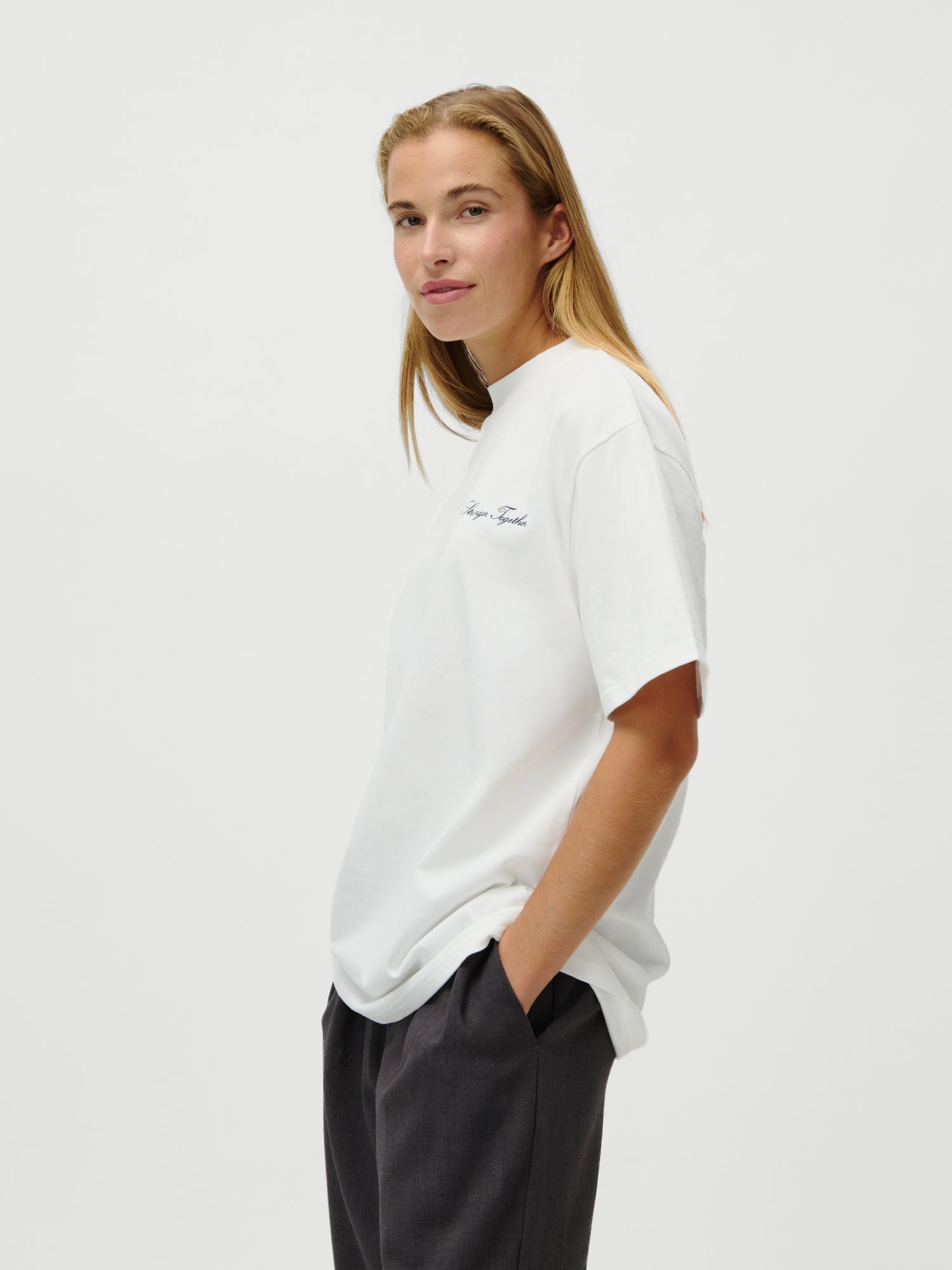 Eine Frau mit langen, hellbraunen Haaren steht vor einem weißen Hintergrund, trägt das LeGer x Oh April - Amara T-Shirt Stronger by LeGer by Lena Gercke und eine dunkle Hose, lächelt leicht und hat die linke Hand in der Tasche.