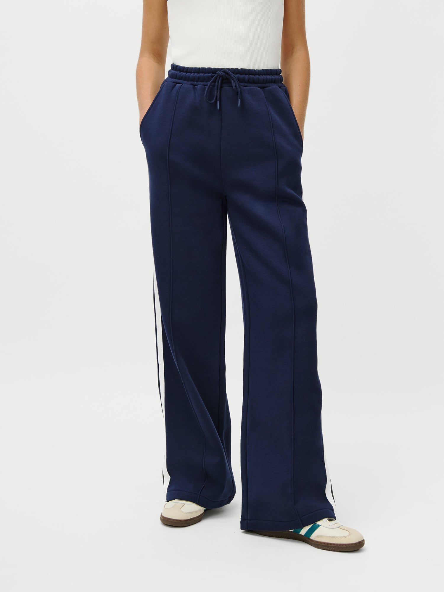 Eine 1,73 m große Person trägt eine marineblaue Jogginghose mit weitem Bein von LeGer x Oh April Joan Jogger von LeGer by Lena Gercke mit weißen Streifen, ein weißes ärmelloses Oberteil und weiße Turnschuhe mit beigen und grünen Akzenten, die Hände in den Taschen.