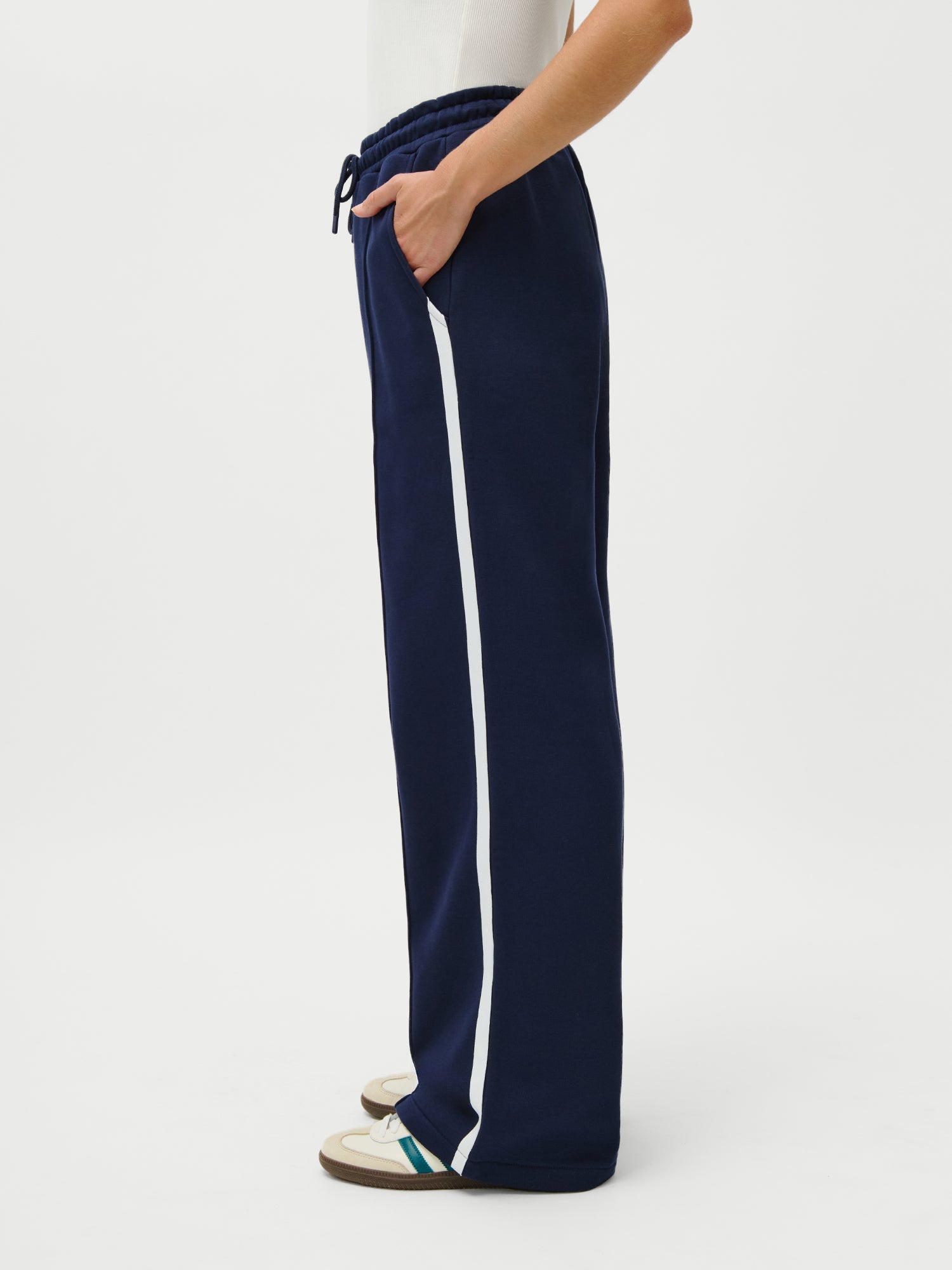 Ein 1,73m großes Model trägt den LeGer x Oh April - Joan Jogger von LeGer by Lena Gercke in navy mit weißem Seitenstreifen, gestylt mit weißen Turnschuhen und einem weißen Oberteil, posiert im Profil vor einem schlichten weißen Hintergrund.