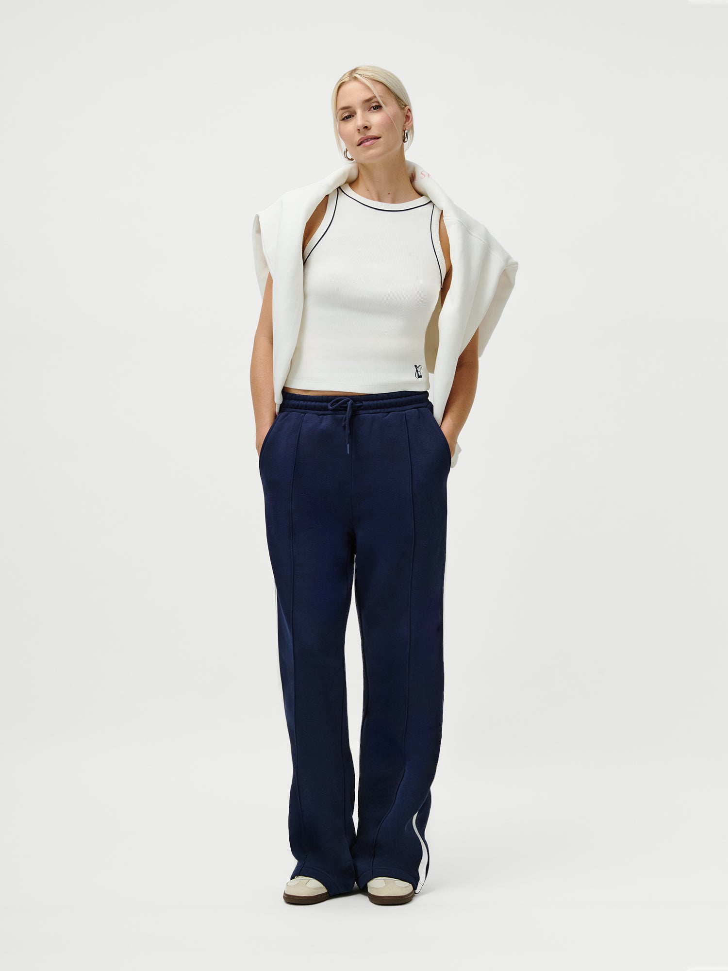 Eine Frau (1,79m, Größe 38) steht vor einem weißen Hintergrund und trägt die marineblaue Hose LeGer x Oh April - Joan Jogger Tall von LeGer by Lena Gercke, gepaart mit einem weißen ärmellosen Oberteil, einem Pullover über den Schultern und weißen Sneakers. Die Hände in den Taschen.