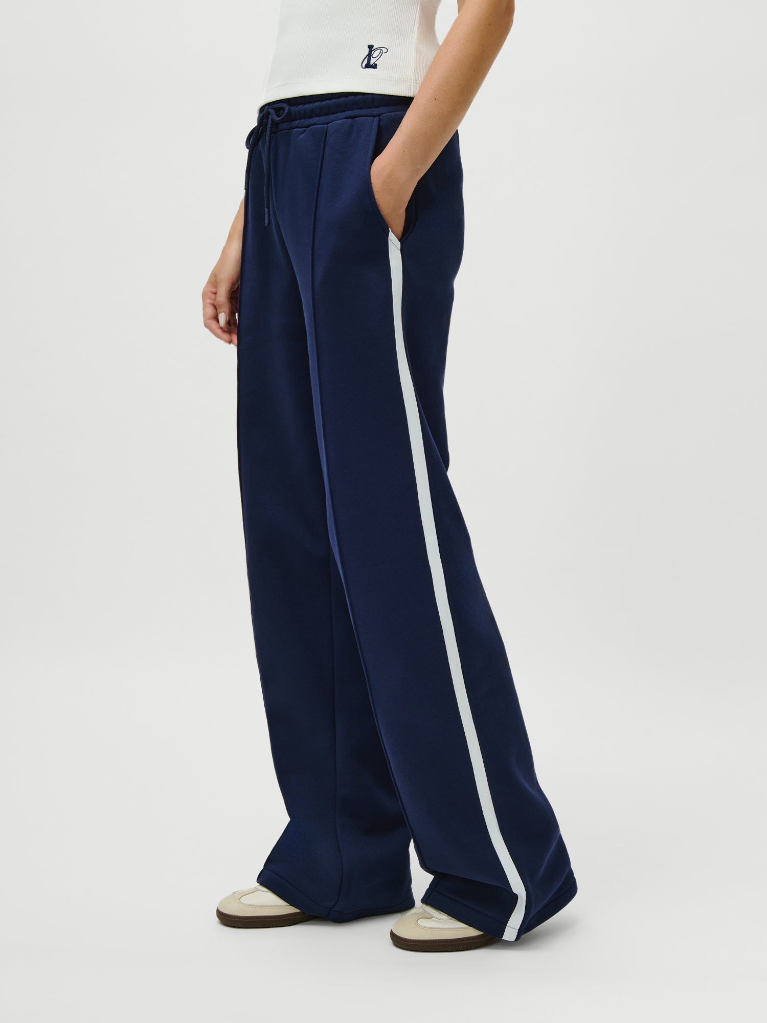 Eine Person (Größe 38, 1,79m) trägt LeGer x Oh April - Joan Jogger Tall von LeGer by Lena Gercke in navy mit weißen Seitenstreifen, gepaart mit einem weißen ärmellosen Oberteil und beigen Turnschuhen, vor einem schlichten weißen Hintergrund.
