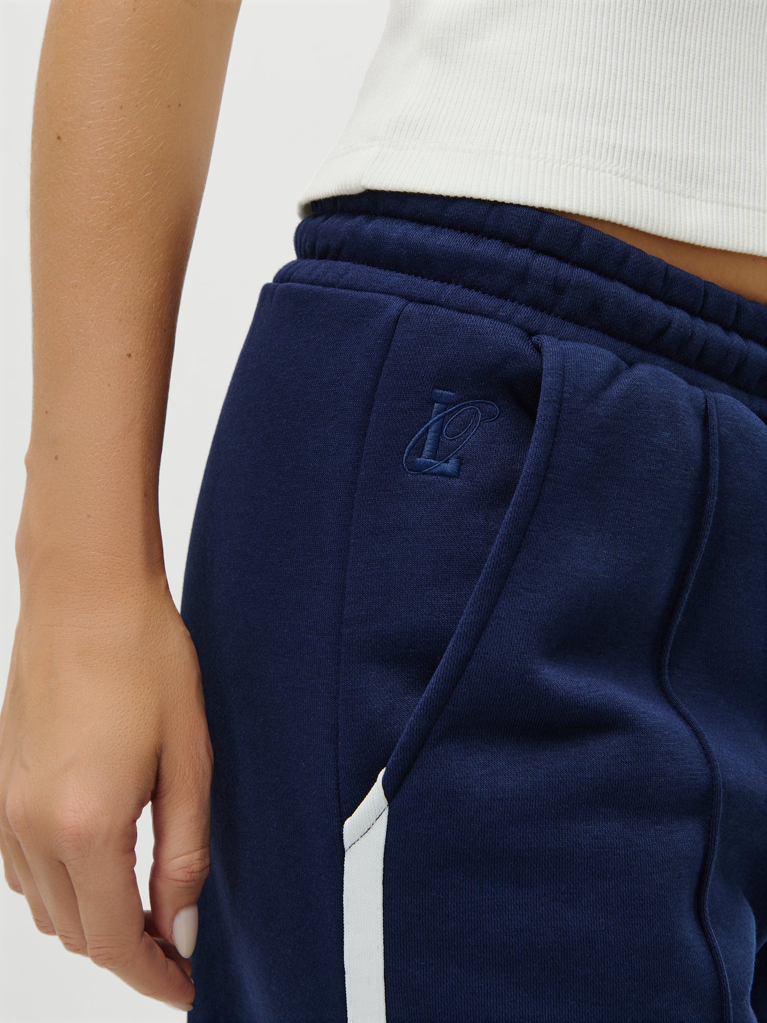 Eine Person trägt die LeGer by Lena Gercke's LeGer x Oh April - Joan Jogger Tall marineblaue Jogginghose mit weißem Streifen (gesticktes Logo neben der Tasche) und ein weißes geripptes ärmelloses Oberteil, Größe 38. Nur der untere Teil des Oberkörpers/der Hand des 1,79m großen Models ist sichtbar.