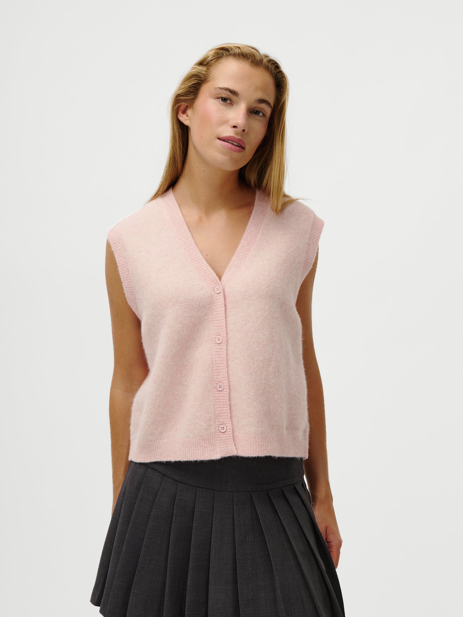 Eine Frau trägt die LeGer x Oh April - Amira Knit Vest von LeGer by Lena Gercke in Hellrosa und einen dunkelgrauen Faltenrock, stehend vor einem weißen Hintergrund. Sie ist 1,73m groß, trägt Größe 36 und blickt entspannt zur Seite.