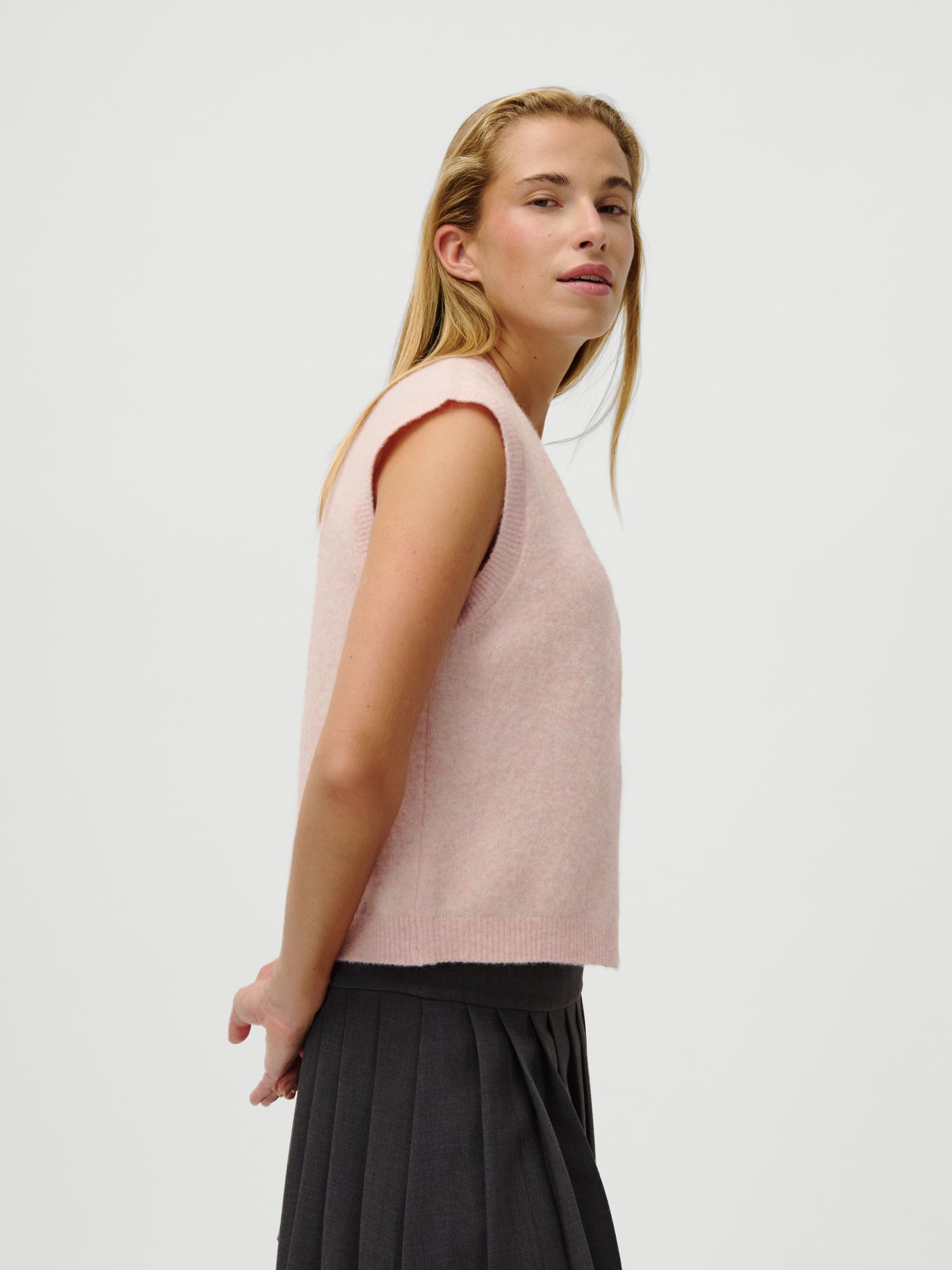 Eine Frau mit langen blonden Haaren trägt die LeGer x Oh April - Amira Knit Vest von LeGer by Lena Gercke in Hellrosa und einen dunklen Faltenrock. Sie steht seitlich und schaut über ihre Schulter vor einem schlichten hellen Hintergrund. Model ist 1,73m, Größe 36, Passform normal.