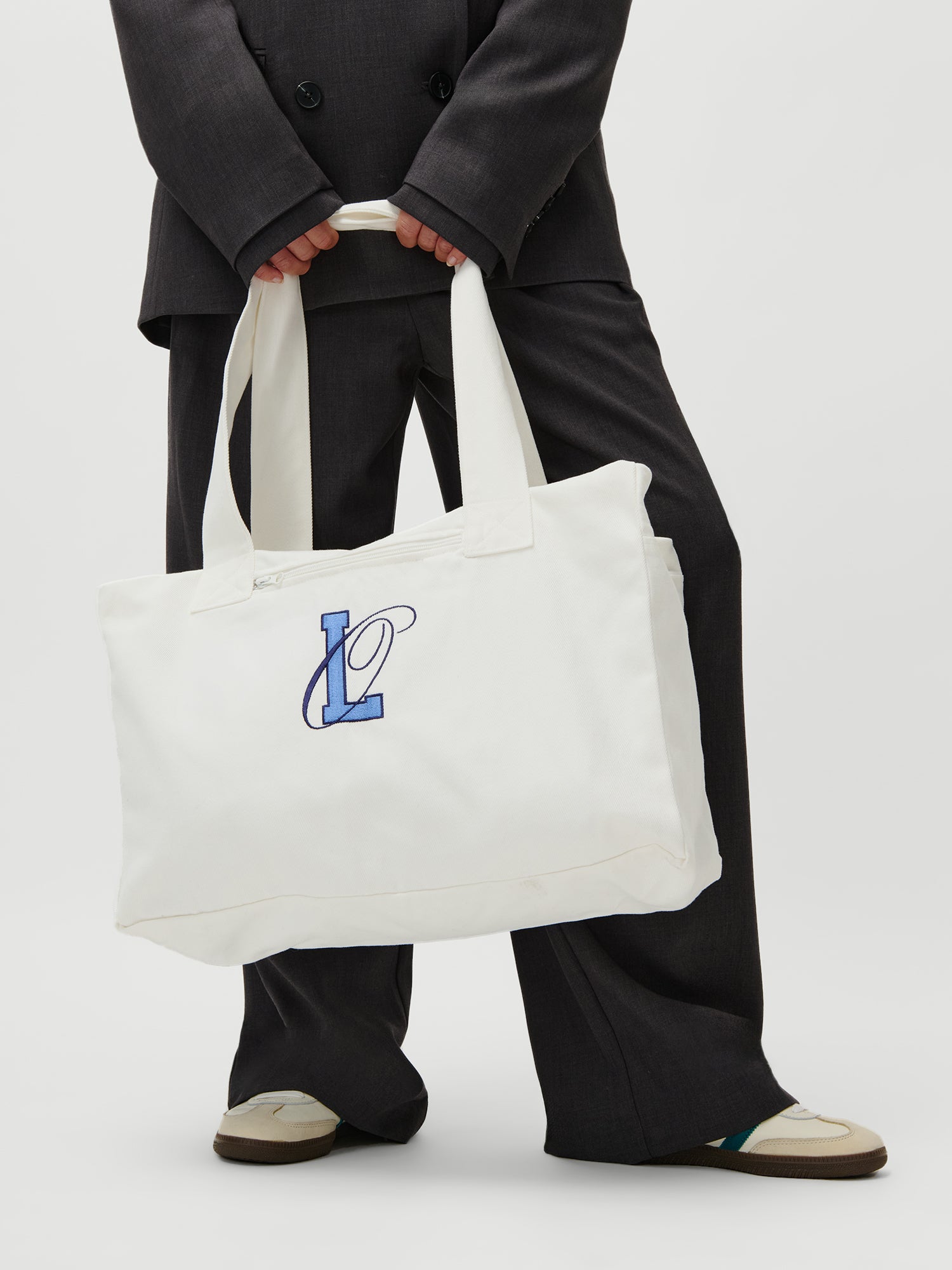 Eine Person in einem dunklen Anzug hält den LeGer x Oh April - Anni Shopper von LeGer by Lena Gercke, eine weiße Canvas-Tasche mit blauer "L"-Stickerei und zwei Henkeln. Das Model ist Größe 36, 1,73m groß und trägt helle Schuhe mit grünen Akzenten vor einem neutralen Hintergrund.