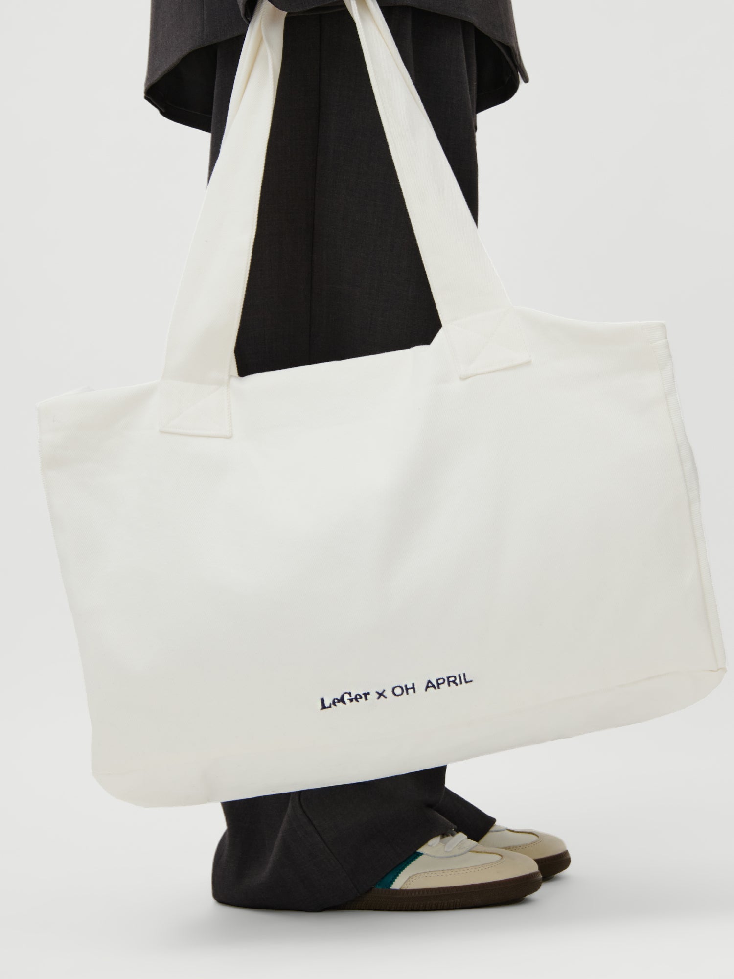 Eine Person in dunkler Hose und Turnschuhen hält den LeGer x Oh April - Anni Shopper von LeGer by Lena Gercke, eine große weiße Tasche mit schwarzer und lila Stickerei. Das Model ist 1,73 m groß und trägt Größe 36.