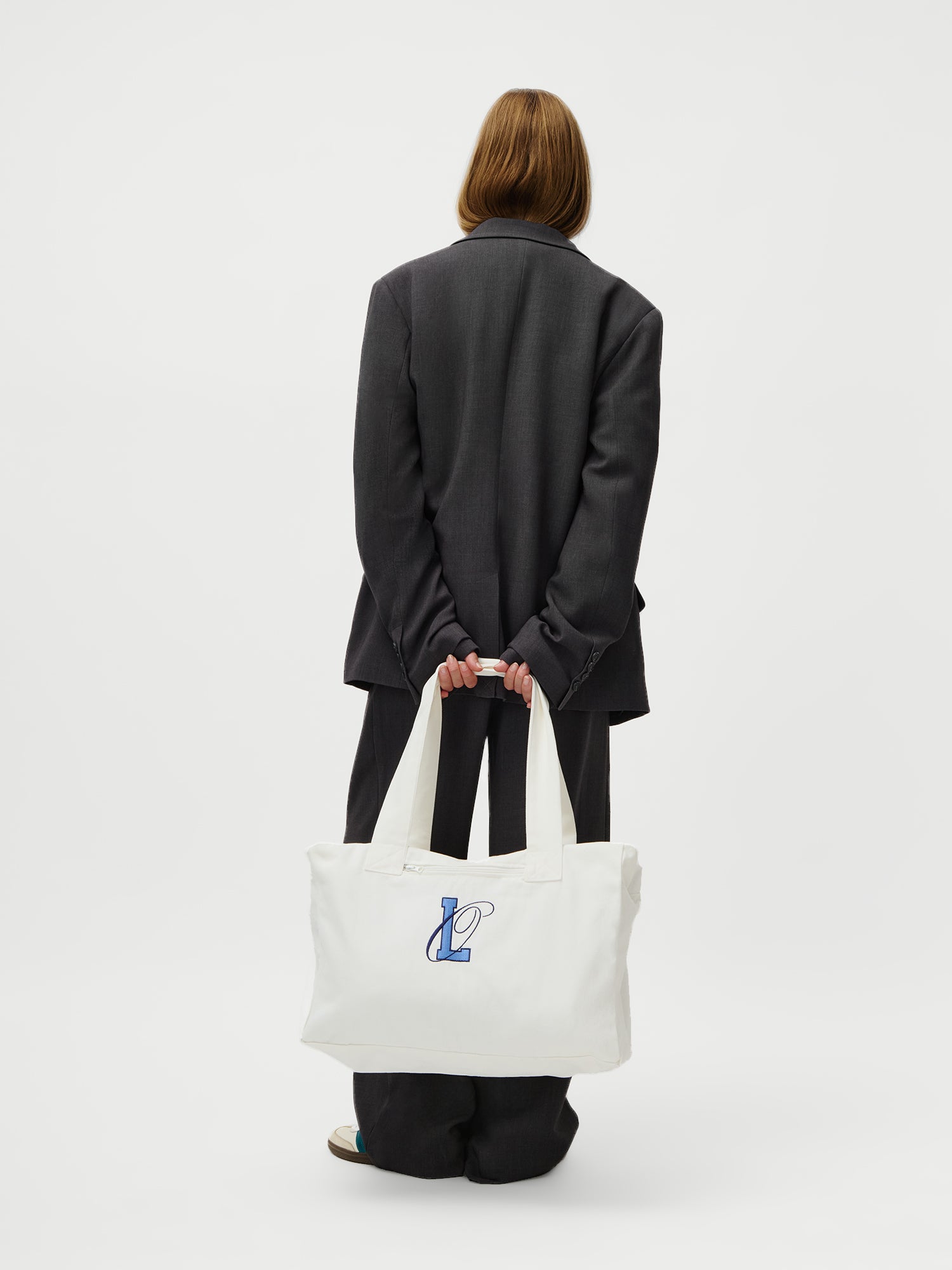 Eine langhaarige Person in einem übergroßen dunklen Anzug (Größe 36), Modelgröße 1,73m, steht mit dem Blick abgewandt und hält den LeGer x Oh April - Anni Shopper von LeGer by Lena Gercke - eine große weiße Tasche mit blauem "L"-Logo - vor einem schlichten weißen Hintergrund.