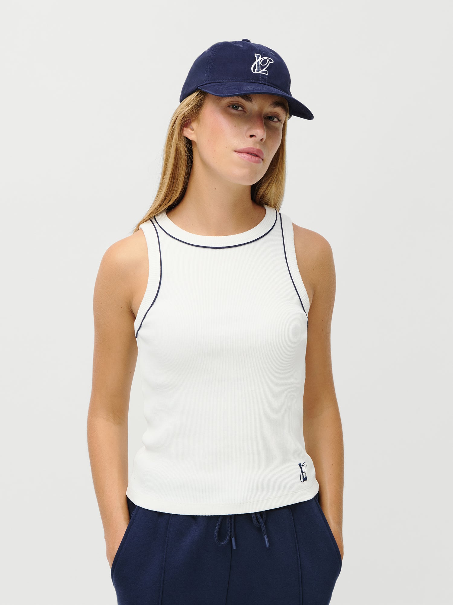 Eine Frau (1,73m, Größe 36) trägt die LeGer x Oh April - Arina Cap Stronger von LeGer by Lena Gercke, ein weißes ärmelloses Oberteil mit marineblauem Saum und eine marineblaue Hose. Sie steht mit den Händen in den Taschen vor einem schlichten weißen Hintergrund.