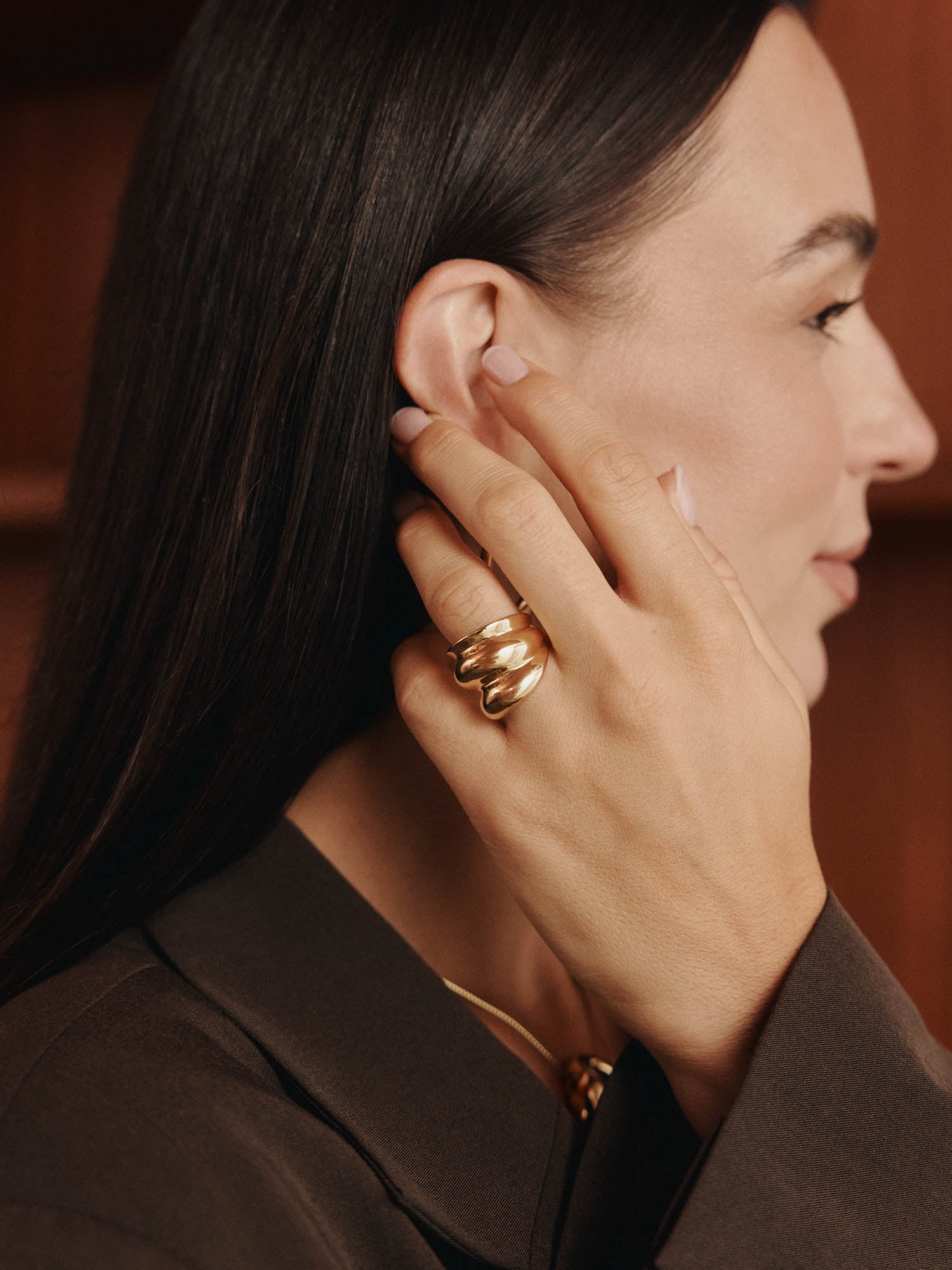 Eine Frau mit langen dunklen Haaren in einer dunklen maßgeschneiderten Jacke berührt ihr Ohr, um den LeGer x Purelei - Dare To Shine Ring von LeGer by Lena Gercke in klobigem Gold hervorzuheben, fotografiert im Profil vor einem warmen braunen Hintergrund.