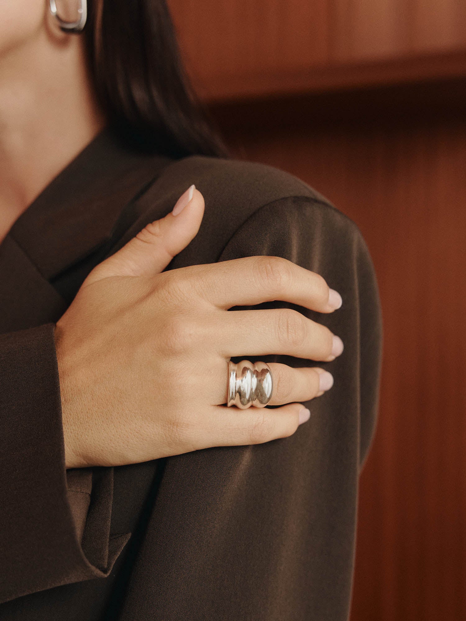 Eine Person in einem braunen Blazer legt ihre Hand auf den Arm und präsentiert den LeGer x Purelei - Dare To Shine Ring von LeGer by Lena Gercke und einen silbernen Creolenring vor einem hölzernen Hintergrund.