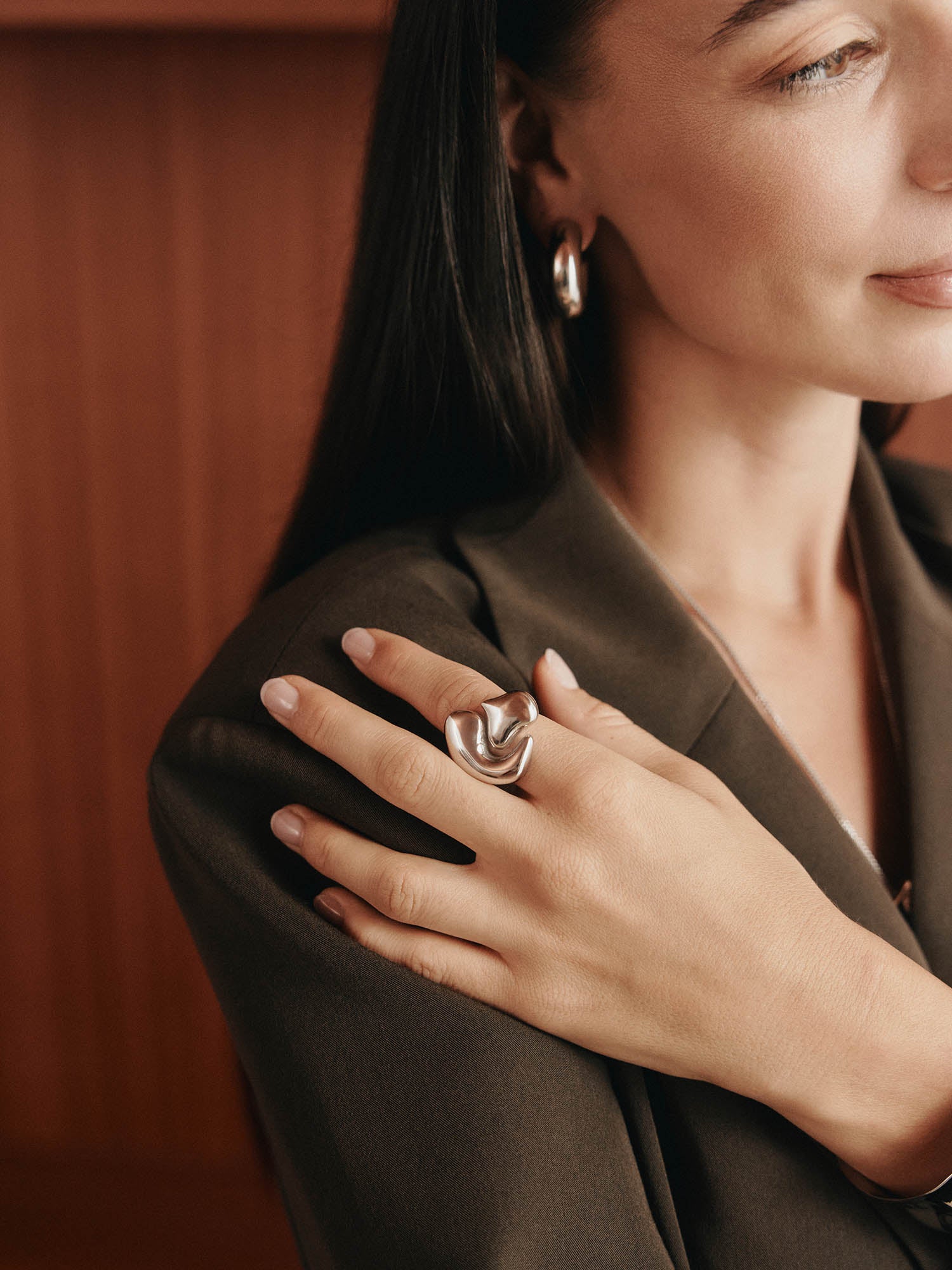 Eine Frau mit langen dunklen Haaren trägt einen braunen Blazer, silberne Ohrringe und den LeGer x Purelei - Fluid Forms Ring von LeGer by Lena Gercke an ihrer Hand, die auf ihrer Schulter vor einem sanft verschwommenen warmtonigen Hintergrund ruht.