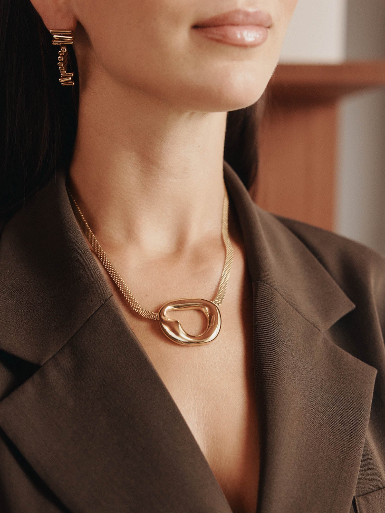 Eine Frau mit dunklem glattem Haar trägt goldene Ohrringe und die LeGer x Purelei - Inner Force Kette von LeGer by Lena Gercke, die einen auffälligen abstrakten Anhänger aufweist. Sie ist von den Schultern aufwärts in einem braunen Blazer mit neutralen Lippen abgebildet.