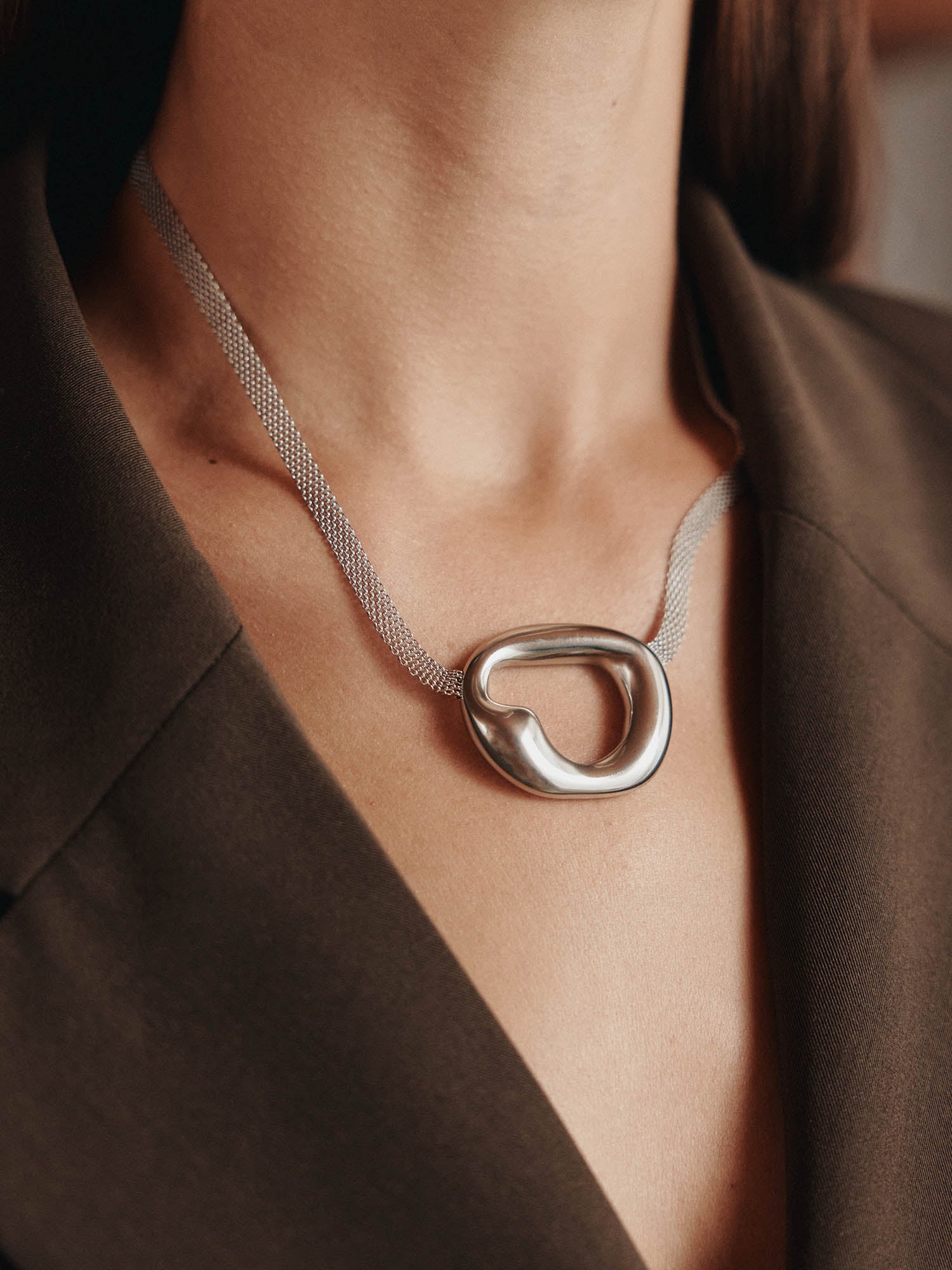 Eine Nahaufnahme einer Person, die einen braunen Blazer und die LeGer x Purelei - Inner Force Kette von LeGer by Lena Gercke trägt - eine auffällige Silberkette mit einem großen, abstrakten, ovalen Anhänger.