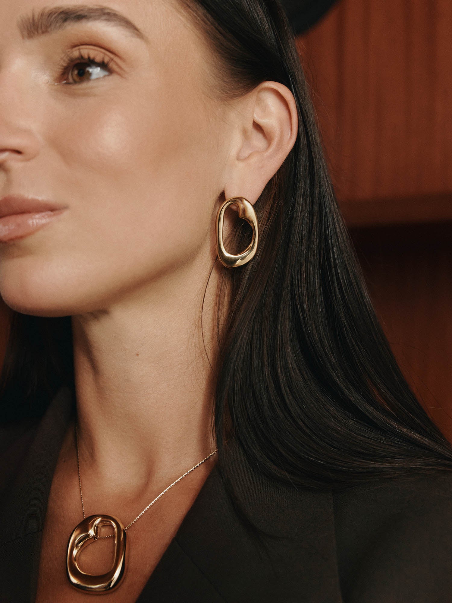 Eine Frau mit glattem dunklem Haar trägt einen braunen Blazer, eine goldene ovale Halskette und LeGer x Purelei - Inner Force Ohrringe von LeGer by Lena Gercke und posiert elegant vor einem Holzhintergrund.