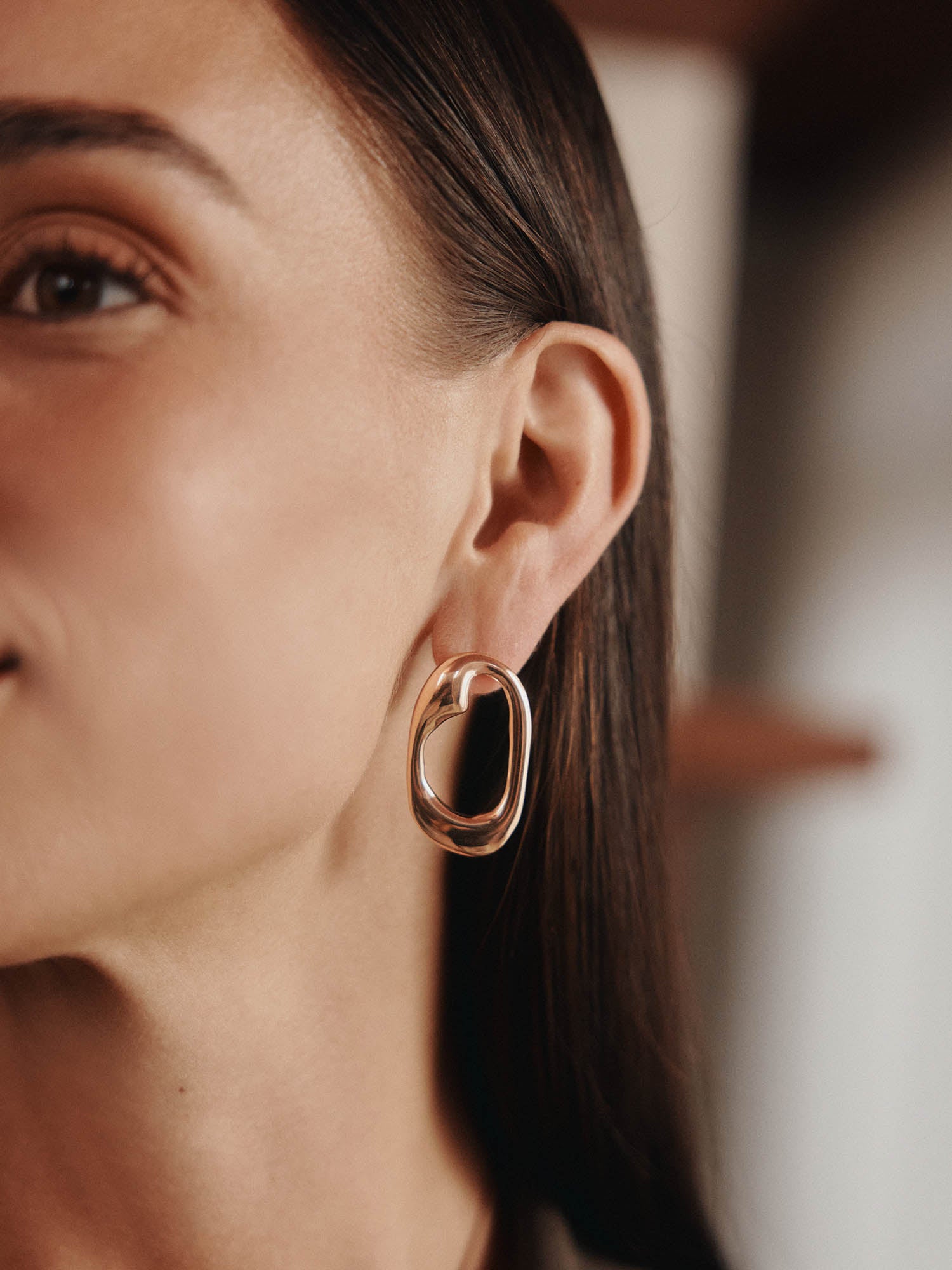 Nahaufnahme eines Frauengesichts, das ihr Ohr hervorhebt, das mit LeGer x Purelei - Inner Force Ohrringe von LeGer by Lena Gercke geschmückt ist. Sie hat glatte Haut, dunkelbraune Haare, die sie hinter ihr Ohr steckt, und einen sanft verschwommenen Hintergrund.