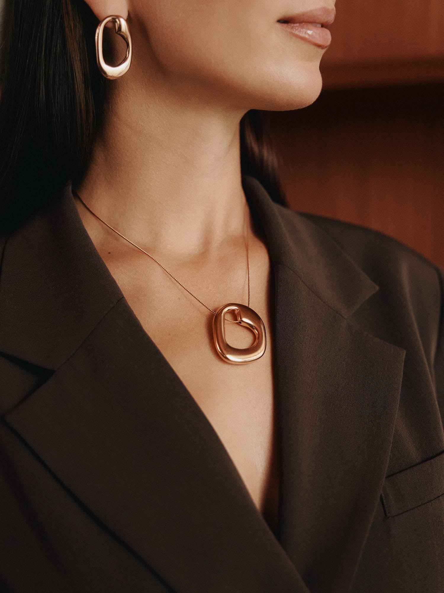 Eine Frau in einem dunkelbraunen Blazer modelliert klobigen Goldschmuck, darunter LeGer x Purelei - Inner Force Ohrringe von LeGer by Lena Gercke und eine große Halskette mit Anhänger. Ihr Gesicht ist teilweise sichtbar, und ihr Haar fällt glatt und locker.