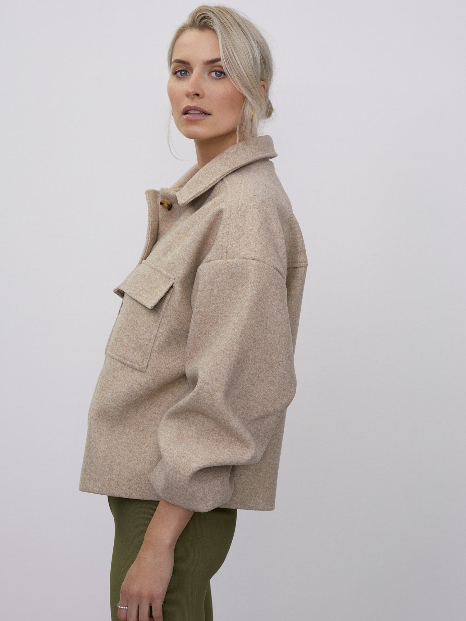 Vor einem schlichten weißen Hintergrund trägt eine blonde Person die Jacke „Eva“ von LeGer by Lena Gercke, eine beige Wolljacke mit großen Taschen und langen Ärmeln, kombiniert mit olivgrünen Hosen. Die lockere Passform verleiht ihr einen lässigen und doch stilvollen Look, da sie leicht zur Seite steht und das Profil der Jacke zur Geltung bringt.