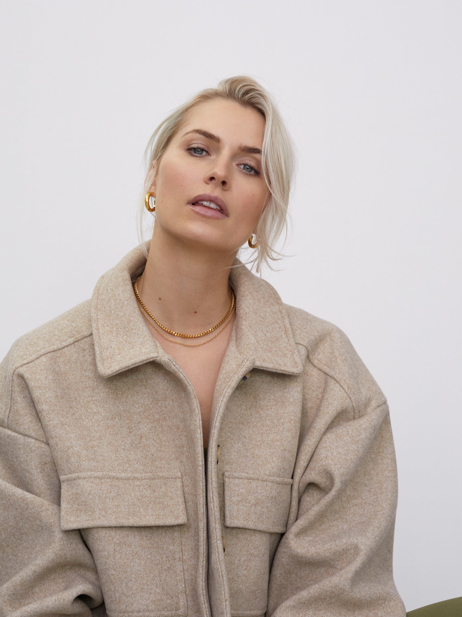 Eine Person mit kurzen, hellblonden Haaren trägt die Jacke „Eva“ von LeGer by Lena Gercke. Diese beige Jacke mit großen Taschen und langem Arm strahlt subtile Eleganz aus. Sie trägt einen neutralen Ausdruck, goldene Creolen und eine einfache goldene Halskette vor einem schlichten weißen Hintergrund.