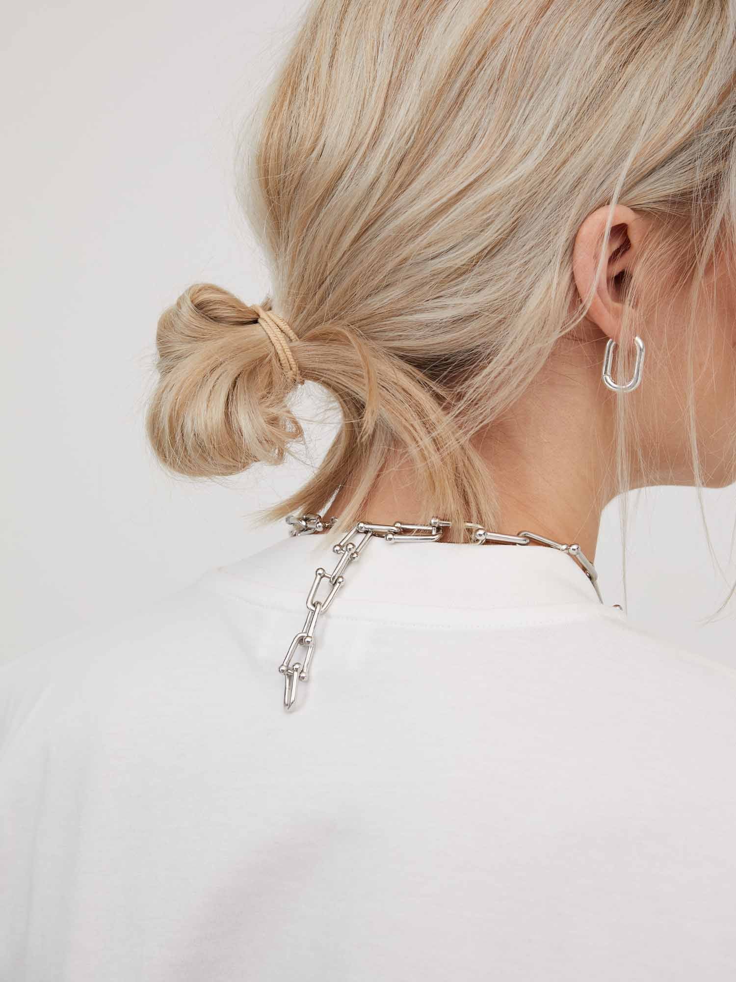 Eine Person mit hellblondem Haar ist von hinten zu sehen. Sie trägt einen tiefen, unordentlichen Dutt, der mit einem Haargummi zusammengehalten wird. Sie trägt ein weißes Hemd „Irina“ von LeGer by Lena Gercke mit Viertelarm-Ärmeln und Accessoires wie eine klobige Silberkette und einen silbernen rechteckigen Creolen-Ohrring. Der Hintergrund ist schlicht und hell.