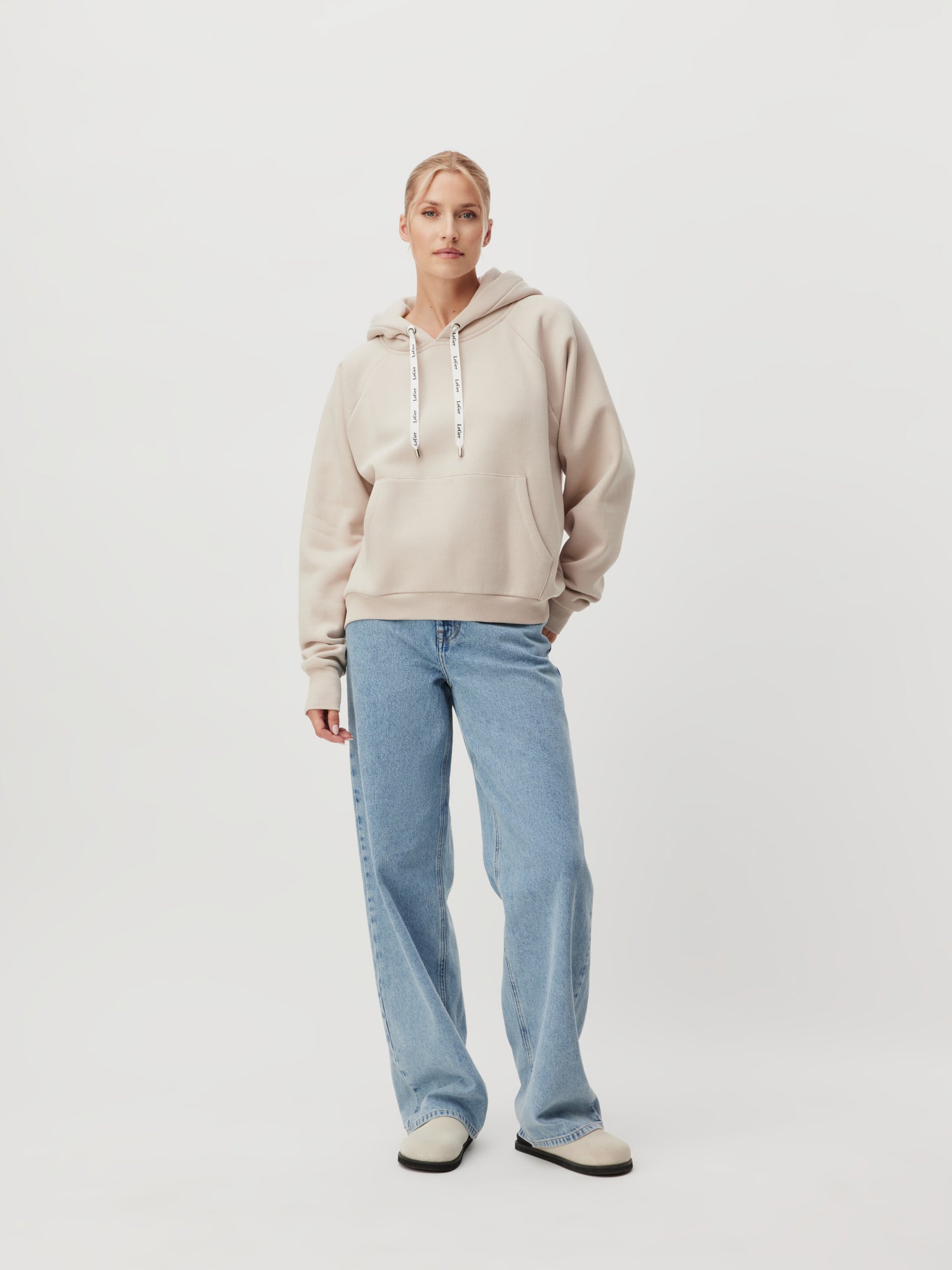 Eine Person steht vor einem schlichten Hintergrund und trägt das hellbeige Sweatshirt „Hayley“ von LeGer by Lena Gercke, dazu hellblaue Jeans und weiße Schuhe. Der langärmlige Hoodie hat Kordeln und eine Vordertasche, die eine perfekte Passform bietet. Die Person hat ihre linke Hand in der Tasche, während die rechte Hand an der Seite hängt.