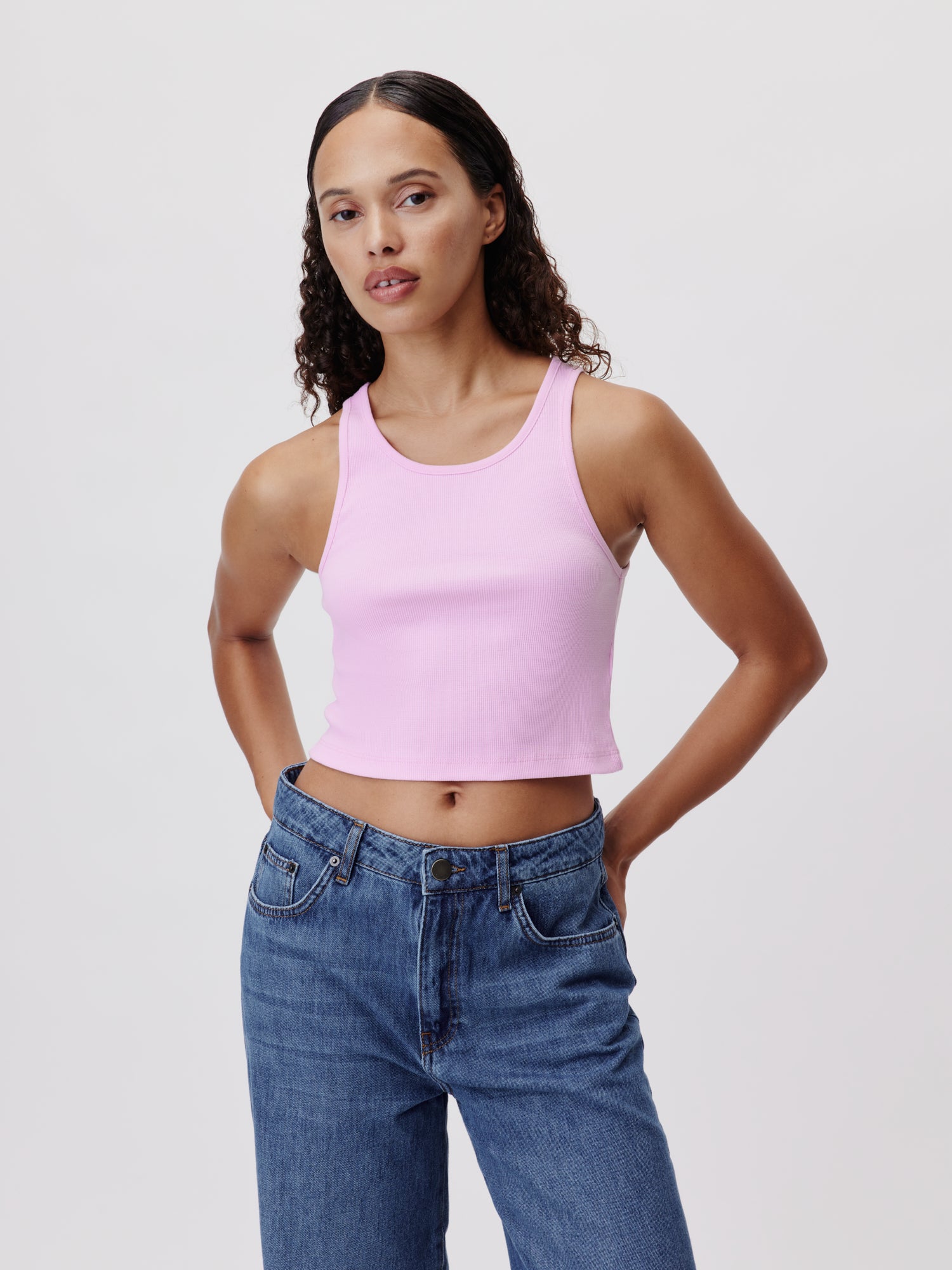 Eine Person mit langen lockigen Haaren trägt das Samantha Top, ein schmales, ärmelloses rosa Crop-Top von LeGer by Lena Gercke, zusammen mit Blue Jeans. Sie steht vor einem schlichten weißen Hintergrund, hat die Hände in die Hüften gestemmt und einen neutralen Gesichtsausdruck.