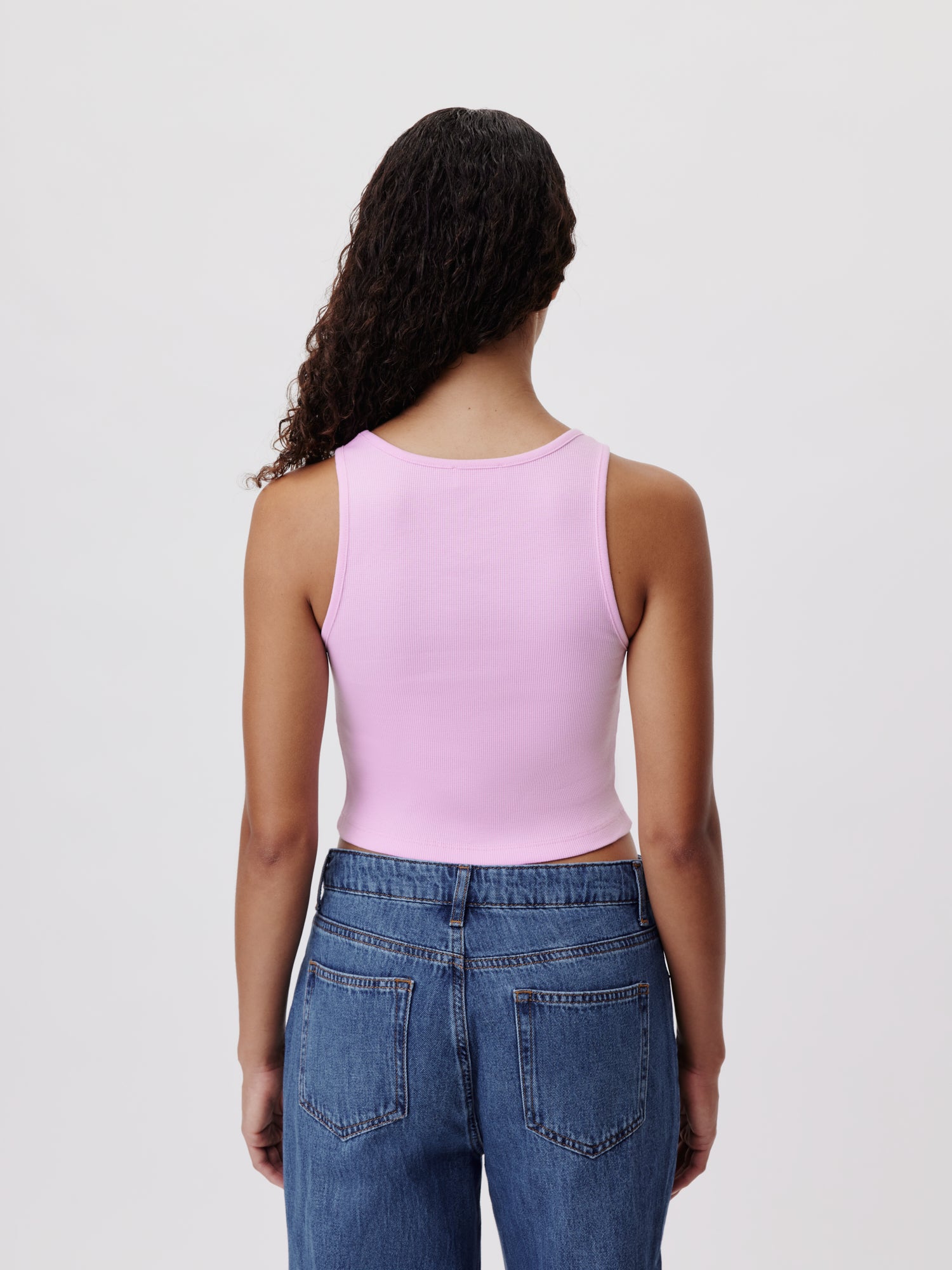 Eine Person mit langen lockigen Haaren trägt ein rosa Samantha Top von LeGer by Lena Gercke im Slim-Design und Blue Jeans. Sie steht mit dem Rücken zur Kamera vor einem schlichten weißen Hintergrund.