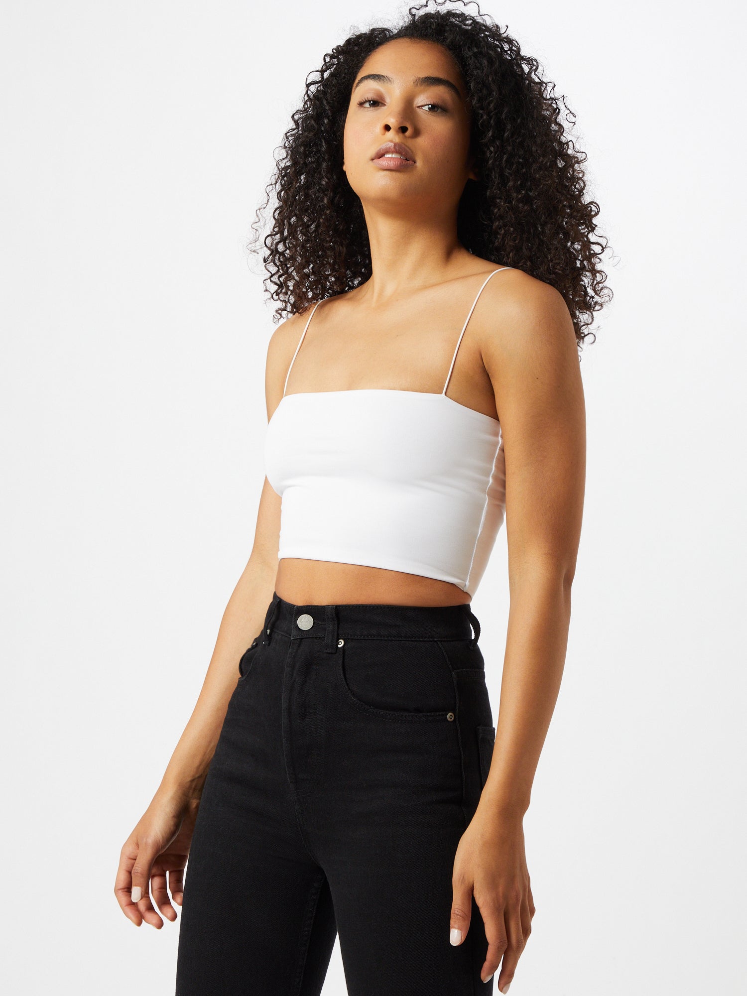 Eine Person mit lockigem Haar steht vor einem schlichten weißen Hintergrund. Sie trägt das figurbetonte, ärmellose weiße „Top Joelle“ von LeGer by Lena Gercke und eine hoch taillierte schwarze Jeans. Sie blickt leicht zur Seite und hat einen neutralen Gesichtsausdruck.