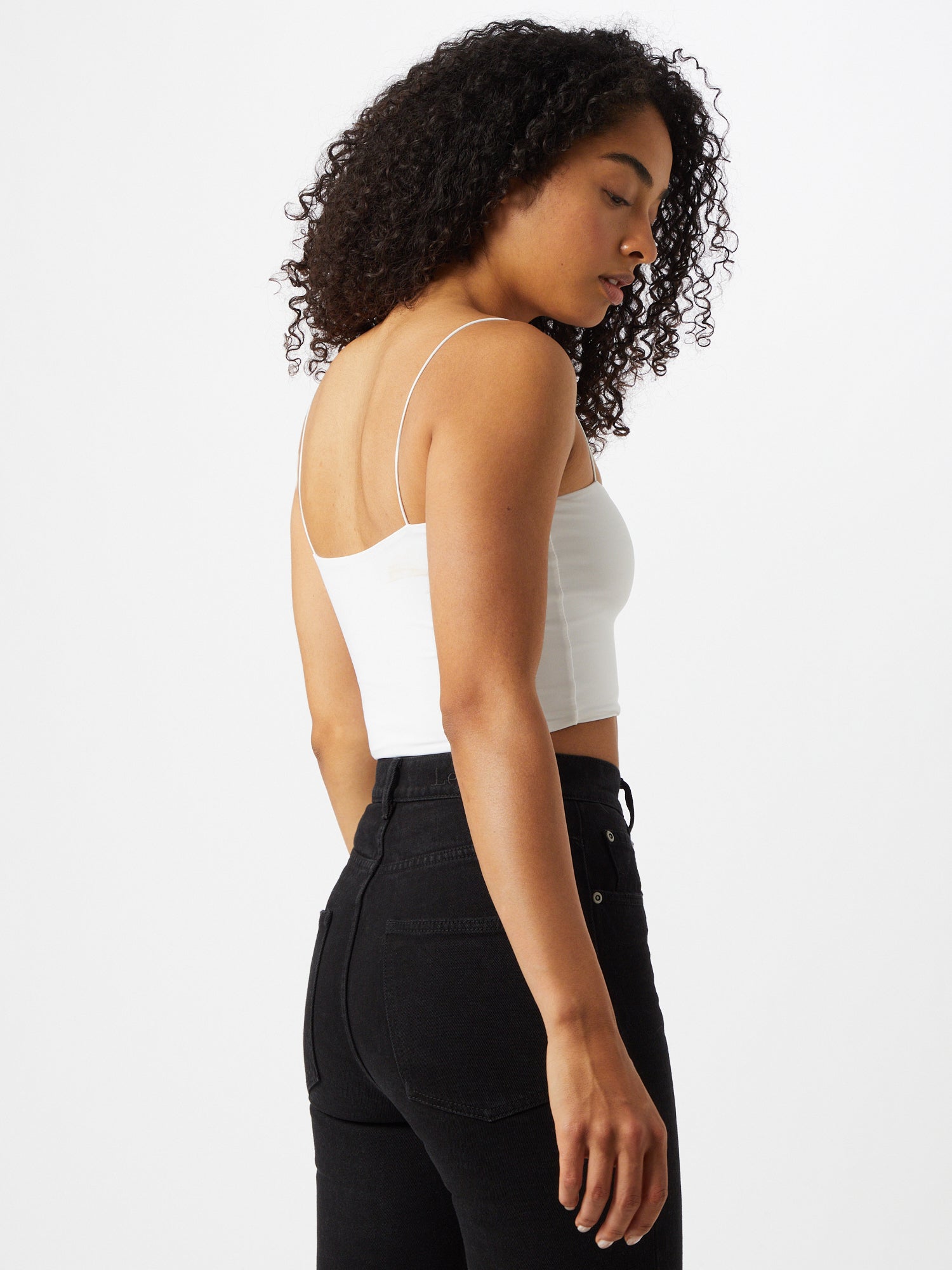 Eine Frau mit lockigem Haar trägt das Top „Joelle“ von LeGer by Lena Gercke, ein weißes Spaghettiträger-Crop-Top, kombiniert mit hoch taillierten schwarzen Jeans. Sie steht seitlich und blickt vor einem schlichten weißen Hintergrund nach unten.