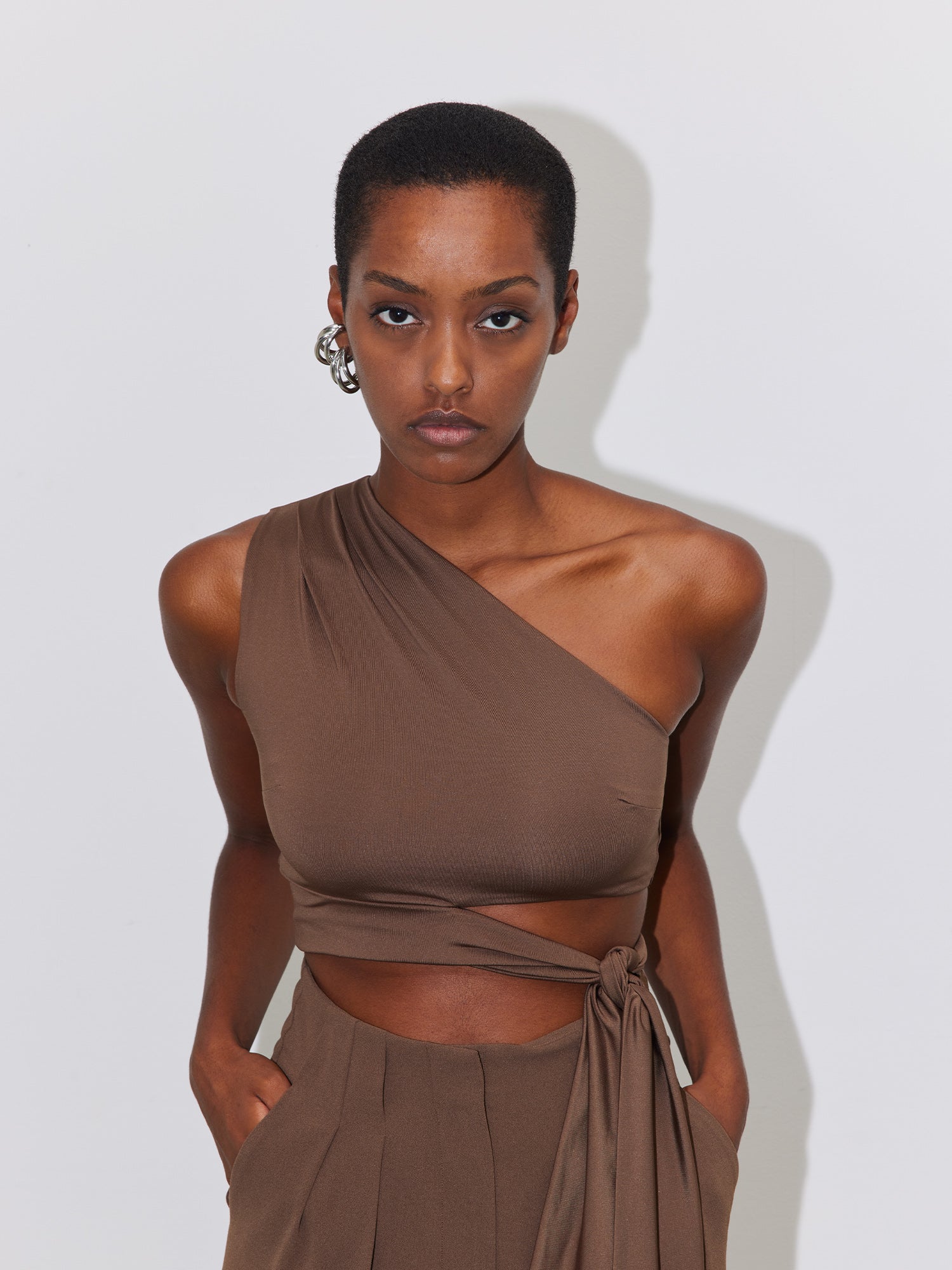 Eine Person mit kurzen Haaren und ernstem Gesichtsausdruck trägt das schulterfreie, ärmellose braune Top „Leia“ von LeGer by Lena Gercke und dazu passende schmale Hosen. Sie hat die Hände in den Taschen und steht vor einem schlichten, hellen Hintergrund. Als Accessoire trägt sie einen Creolenohrring.