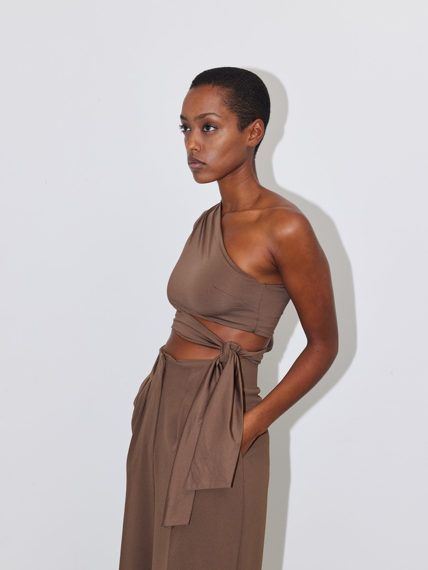 Eine Frau mit Kurzhaarschnitt steht vor einem schlichten weißen Hintergrund. Sie trägt das stylische, ärmellose One-Shoulder-Top „Leia“ von LeGer by Lena Gercke mit Wickeldetail und dazu passende High-Waist-Hosen. Sie hat die Hände in den Taschen und blickt zur Seite.