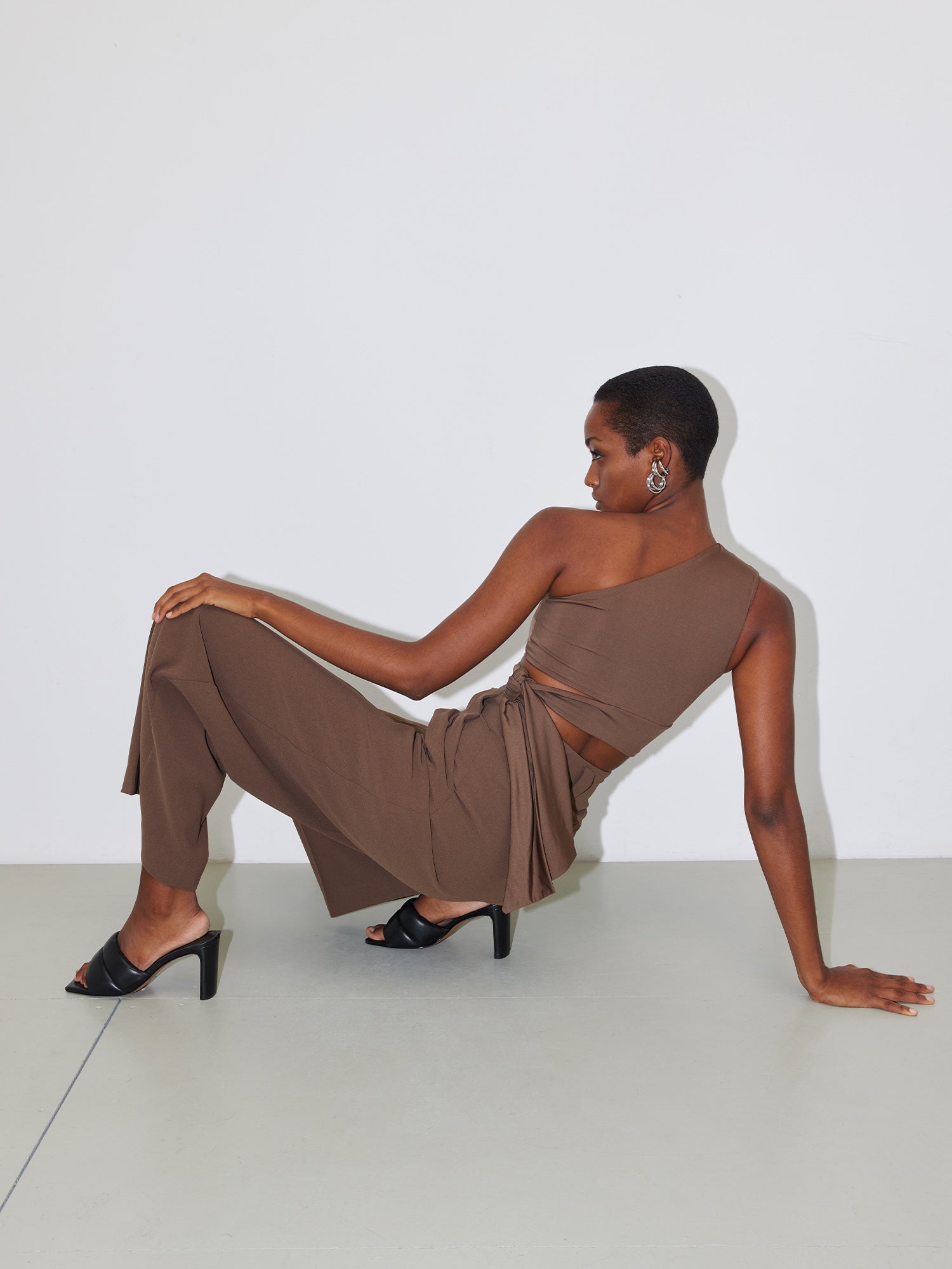 Eine Person posiert stilvoll vor einem schlichten weißen Hintergrund. Sie ist mit einem braunen, schmalen Top „Leia“ von LeGer by Lena Gercke, einer passenden Hose und schwarzen High Heels bekleidet. Die Pose besteht aus einem nach hinten gestreckten Arm, der die sitzende Position stützt, während der andere Arm auf dem angehobenen Knie ruht.