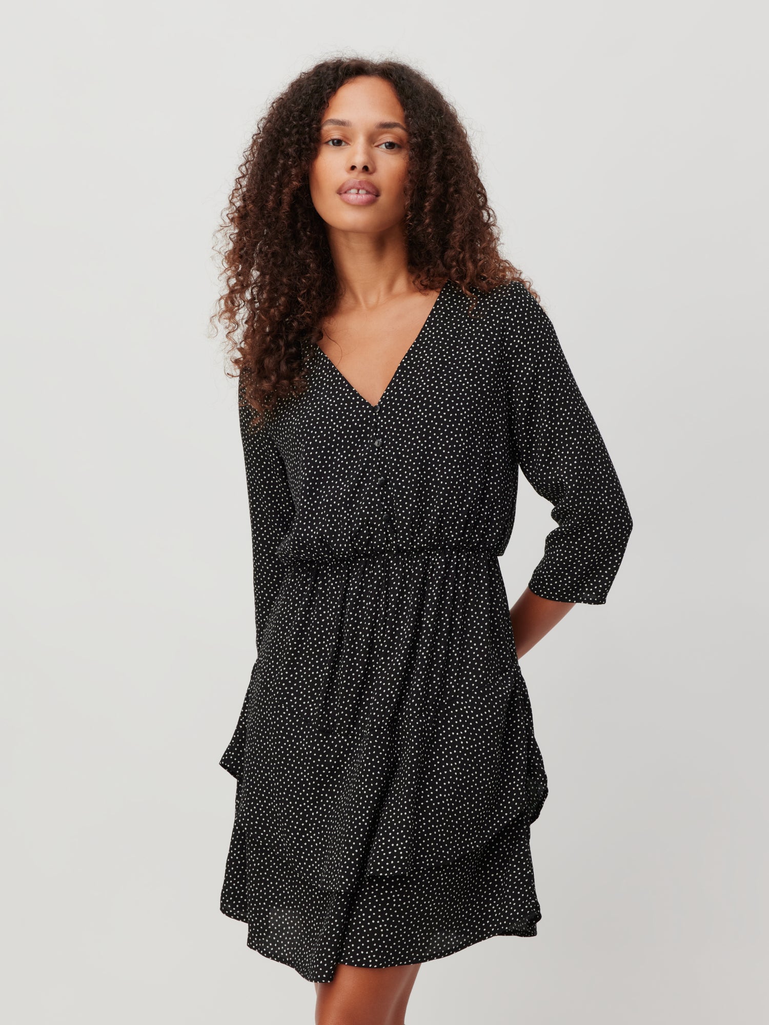 Jemand mit lockigem Haar trägt das Mara Dress von LeGer by Lena Gercke, ein schwarzes Minikleid, das mit winzigen weißen Punkten verziert ist. Dieses schicke Outfit verfügt über einen V-Ausschnitt, einen elastischen Bund und Dreiviertelärmel. Der schlichte cremefarbene Hintergrund ergänzt es perfekt.