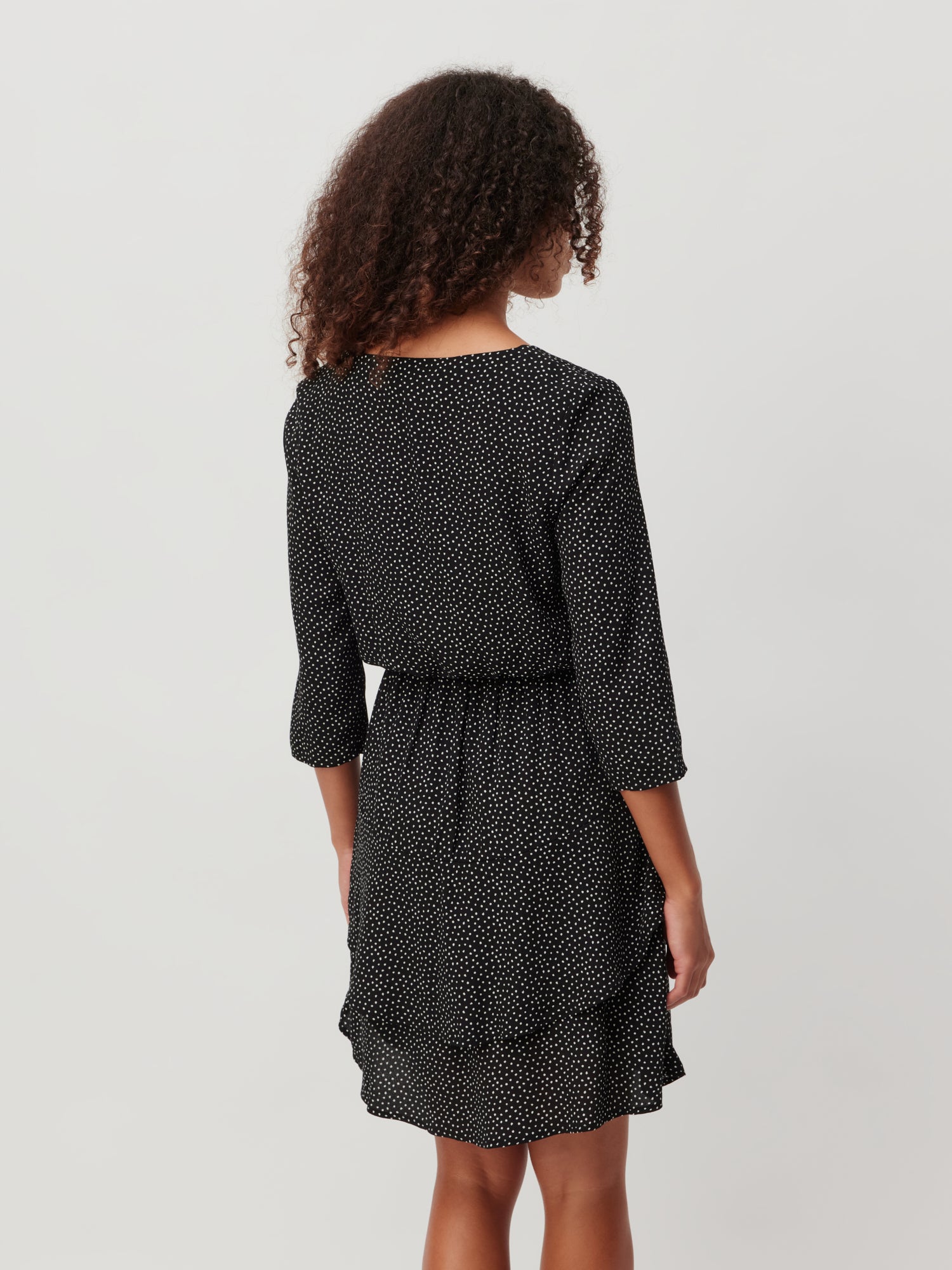 Eine Person mit lockigem Haar steht abgewandt und hebt das Mara-Kleid von LeGer by Lena Gercke hervor. Dieses schwarze Kleid von Passform mit weißen Punkten hat Dreiviertelärmel, eine taillierte Taille und einen Lagenrock vor einem schlichten hellgrauen Hintergrund.