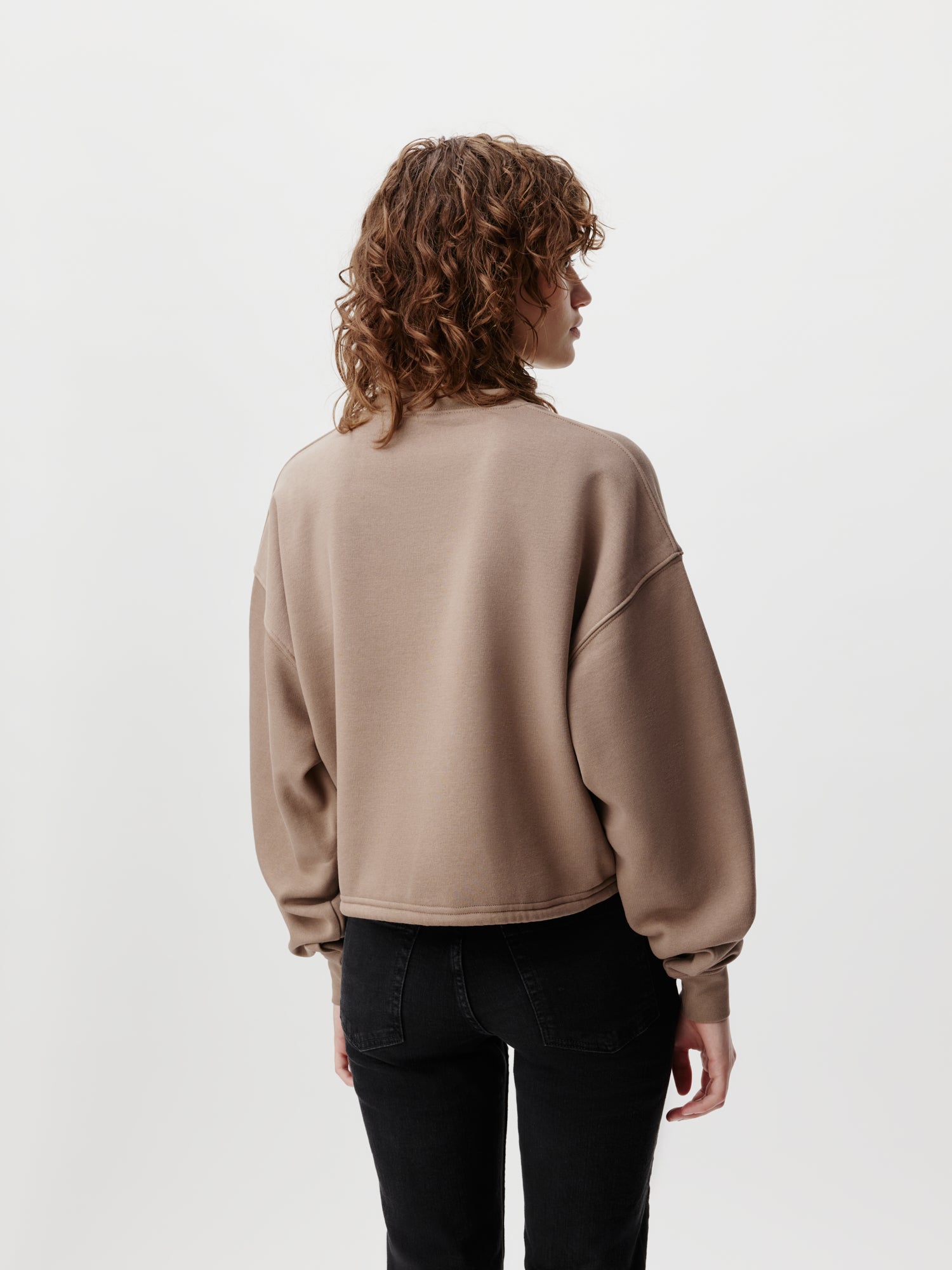 Eine Person mit lockigem Haar trägt ein taupefarbenes Sweatshirt „Rosa“ von LeGer by Lena Gercke und schwarze Jeans und steht mit dem Rücken zu einem schlichten weißen Hintergrund.