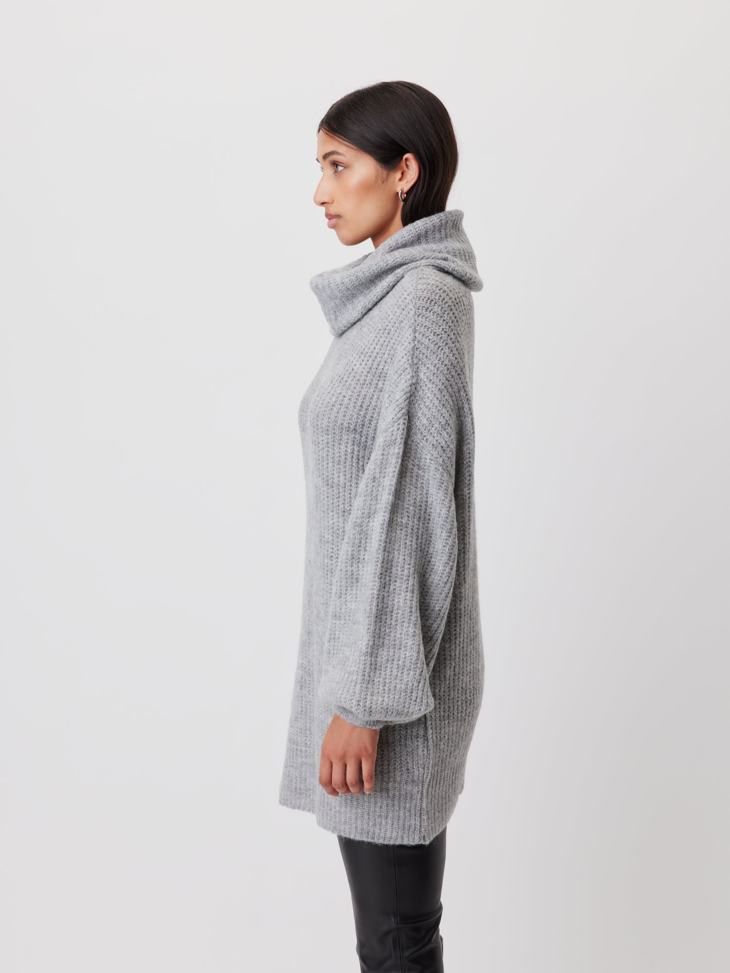 Eine Person mit dunklem Haar steht im Profil vor einem schlichten Hintergrund. Sie trägt einen gemütlichen, übergroßen grauen Pullover „Juna“ von LeGer by Lena Gercke mit großem Wasserfallausschnitt und schwarzen Hosen. Der lange Schnitt des Pullovers unterstreicht seine entspannte Passform. Ihr Gesichtsausdruck ist neutral und ihre Hände ruhen locker an ihren Seiten.