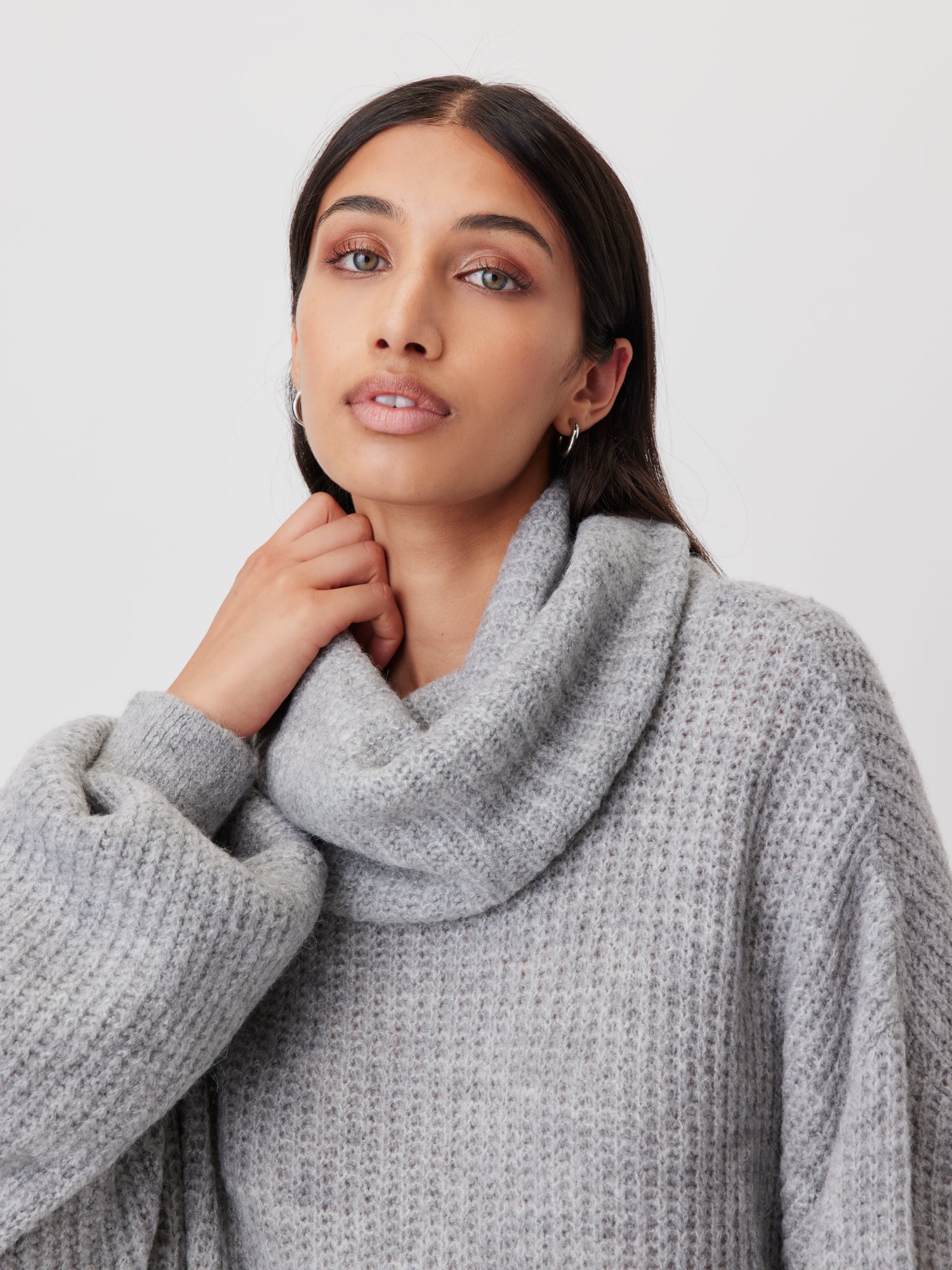 Eine Person mit langen, dunklen Haaren trägt den gemütlichen, hellgrauen Pullover „Juna“ von LeGer by Lena Gercke. Der Pullover hat einen Wasserfallausschnitt und eine etwas längere Passform, die den Tragekomfort erhöht. Sie hat einen neutralen Gesichtsausdruck und berührt sanft mit einer Hand den Kragen ihres Pullovers vor einem schlichten weißen Hintergrund.