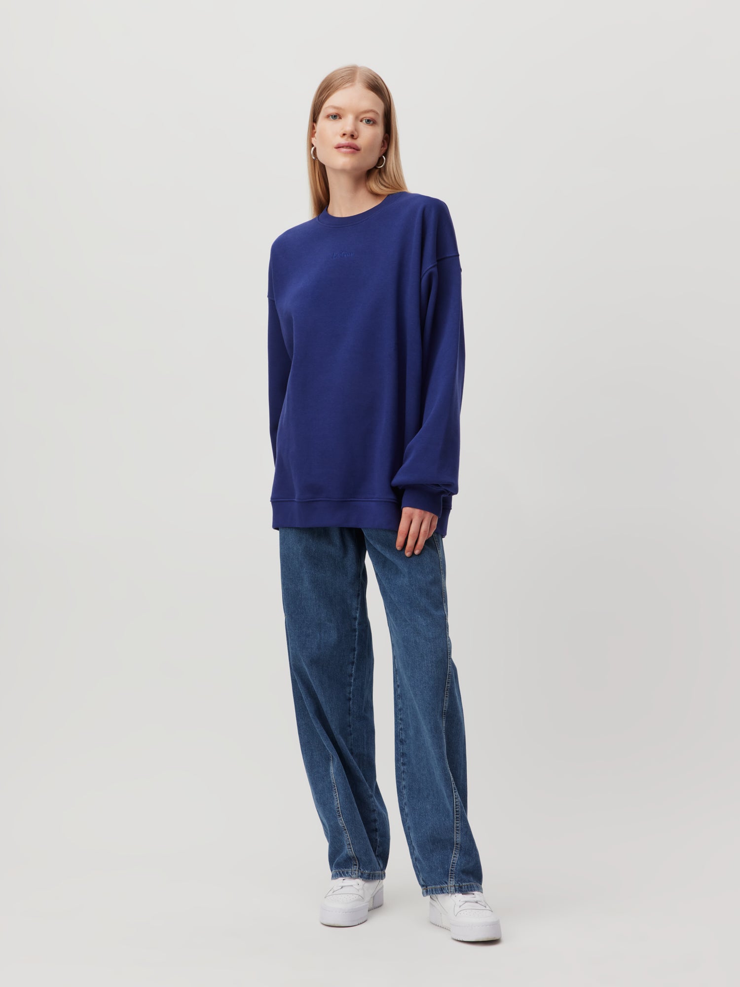 Eine Person mit langen Haaren steht vor einem schlichten Hintergrund und trägt ein locker sitzendes marineblaues Sweatshirt „Indra“ von LeGer by Lena Gercke und hellblaue Jeans, ergänzt durch weiße Sneakers. Die Pose ist entspannt mit einer Hand in der Tasche.