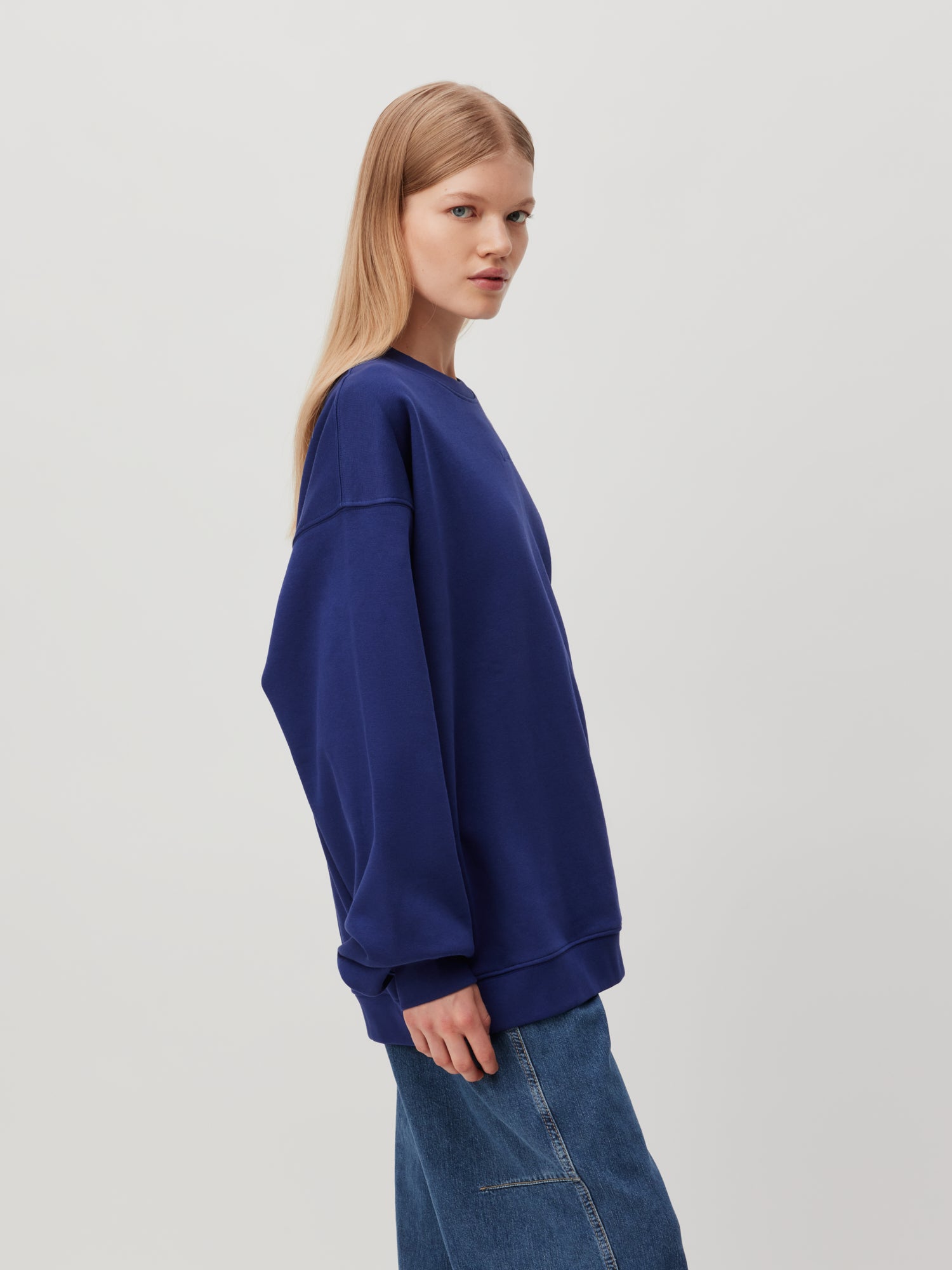 Eine Person mit langen, glatten blonden Haaren trägt ein locker sitzendes, übergroßes dunkelblaues Sweatshirt „Indra“ von LeGer by Lena Gercke und dunkelblaue Jeans. Sie steht seitlich, blickt leicht nach links und hat einen neutralen Gesichtsausdruck vor einem schlichten hellgrauen Hintergrund.