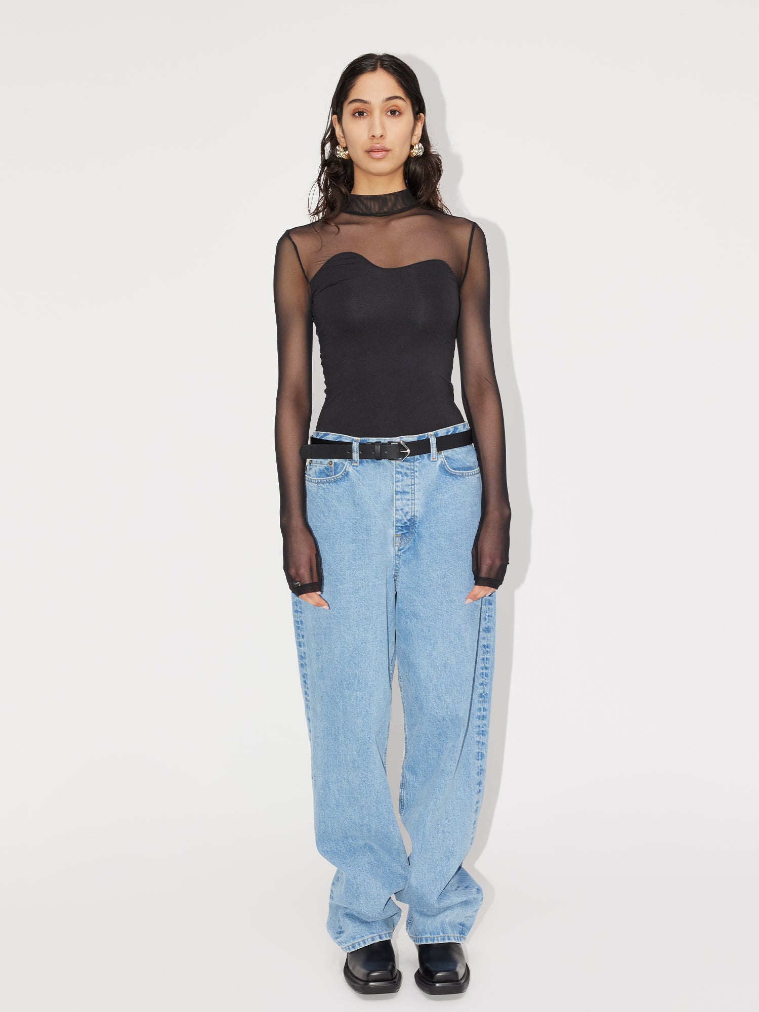Eine Person mit langen, dunklen Haaren steht vor einem weißen Hintergrund und trägt das schwarze Mesh-Shirt „Auguste“ von LeGer by Lena Gercke für einen eleganten Schnitt. Ihre blauen Baggy-Jeans haben eine normale Länge, dazu passen schwarze Schuhe und ihre Hände hängen entspannt an den Seiten.