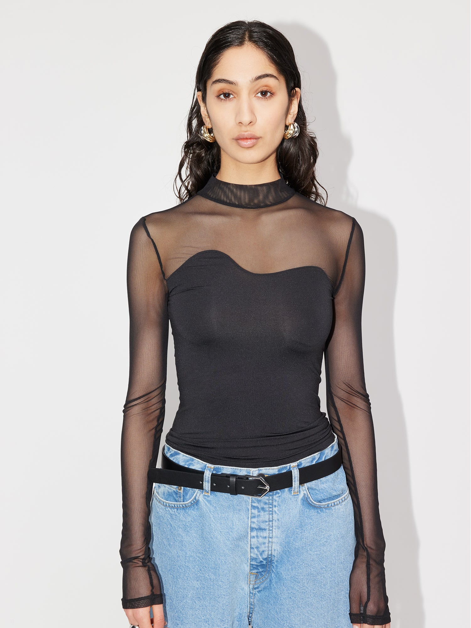 Eine Frau mit langen dunklen Haaren trägt das transparente schwarze Langarm-Oberteil „Auguste“ von LeGer by Lena Gercke, das seine normale Länge und elegante Passform betont, über einem trägerlosen Oberteil. Sie kombiniert es mit hellblauen Jeans und einem schwarzen Gürtel vor einem makellos weißen Hintergrund.