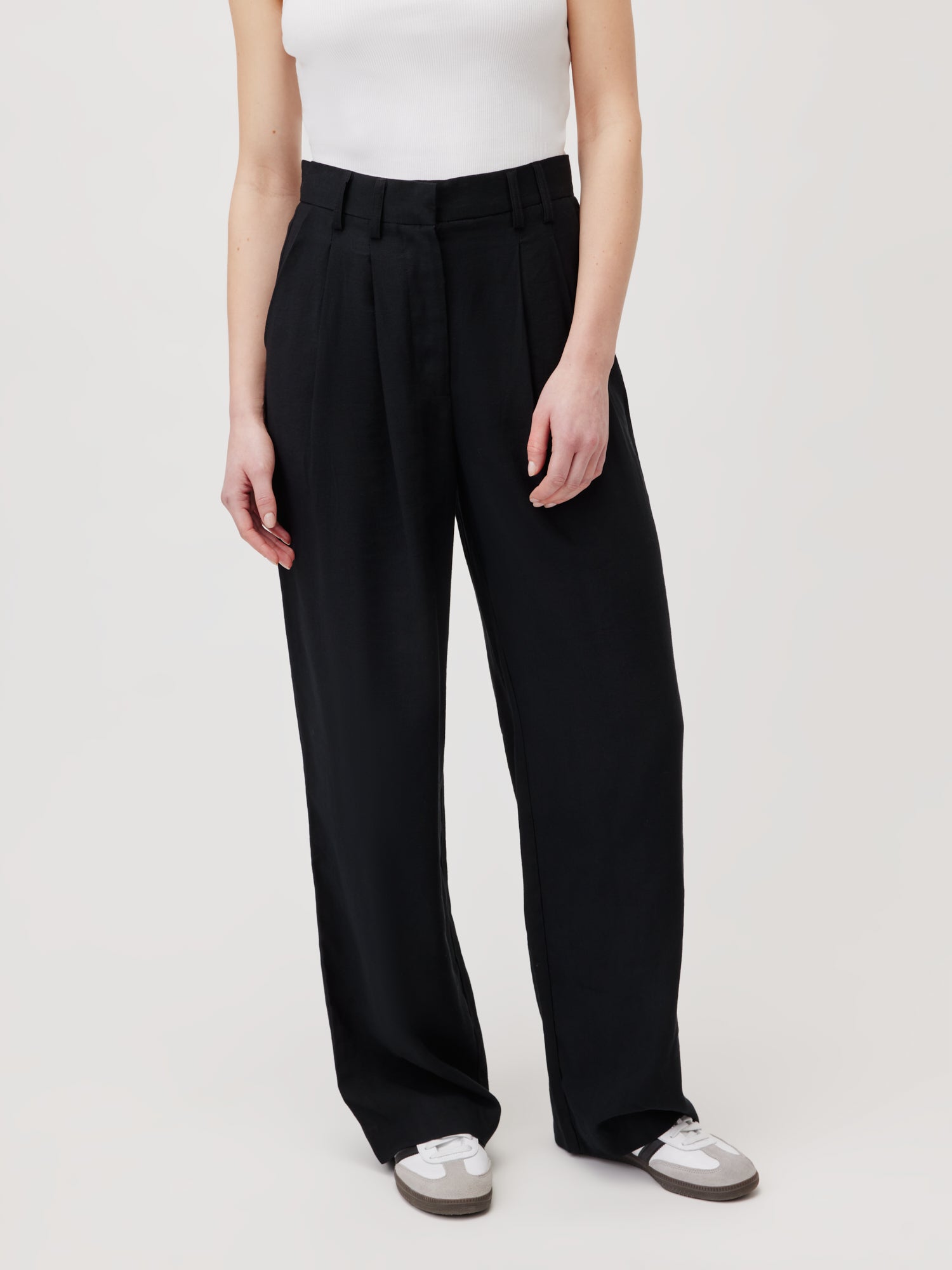 Die Person trägt eine hoch taillierte, schwarze Hose mit weitem Bein namens „Hose Draco“ von LeGer by Lena Gercke und ein weißes ärmelloses Oberteil mit lockerer Passform. Den Look komplettieren sie mit weißen und grauen Sneakers vor einem schlichten, hellen Hintergrund.