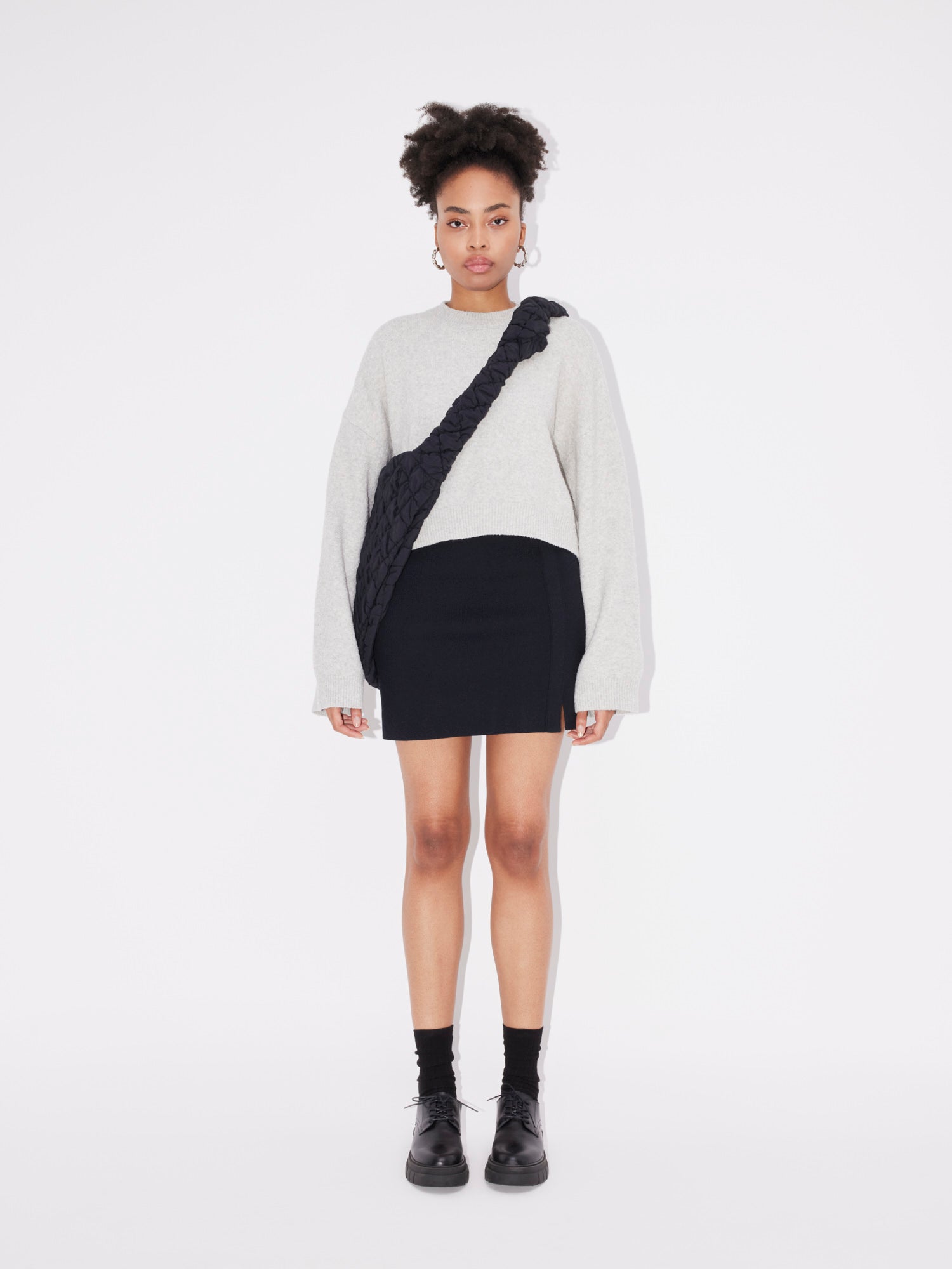 Eine Person steht vor einem weißen Hintergrund und trägt den Pullover „Elwine“ von LeGer by Lena Gercke, einen hellgrauen Langarmpullover mit lockerer Passform, dazu einen schwarzen Minirock, schwarze Socken und schwarze Schuhe. Als Accessoires trägt sie außerdem eine schwarze Umhängetasche mit strukturiertem Riemen.