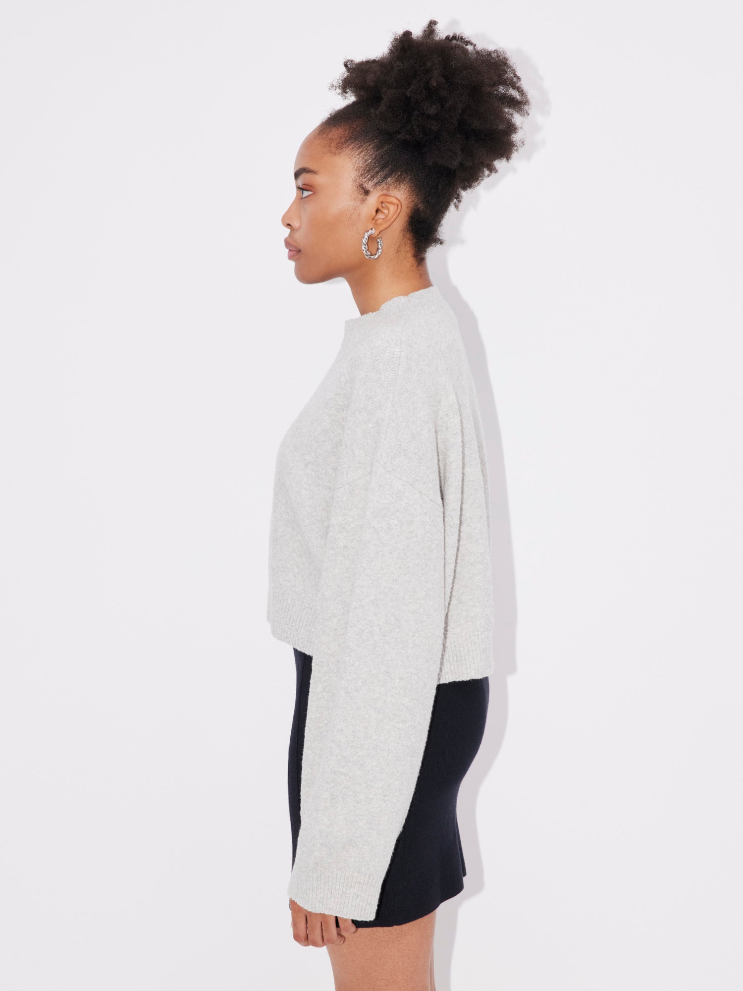 Eine Person mit Afrofrisur steht im Profil und trägt den hellgrauen Langarmpullover „Elwine“ von LeGer by Lena Gercke und einen schwarzen Rock. Ihr Outfit wirkt wie ein Spind und silberne Creolen vervollständigen den Look vor einem schlichten weißen Hintergrund.