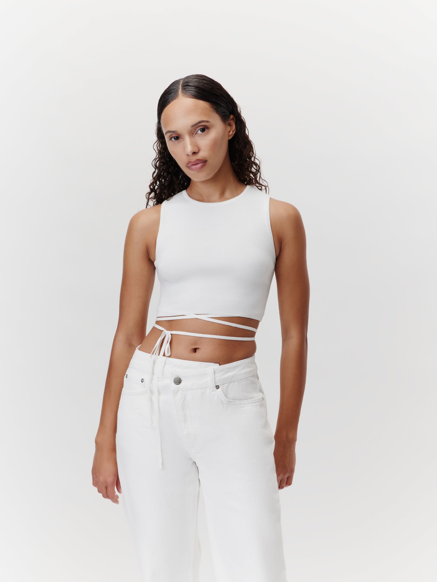 Eine Person mit langen lockigen Haaren trägt das Leany Top, ein weißes kurzes Design mit Wickelbändern von LeGer by Lena Gercke, kombiniert mit figurbetonten weißen Jeans vor einem schlichten weißen Hintergrund.
