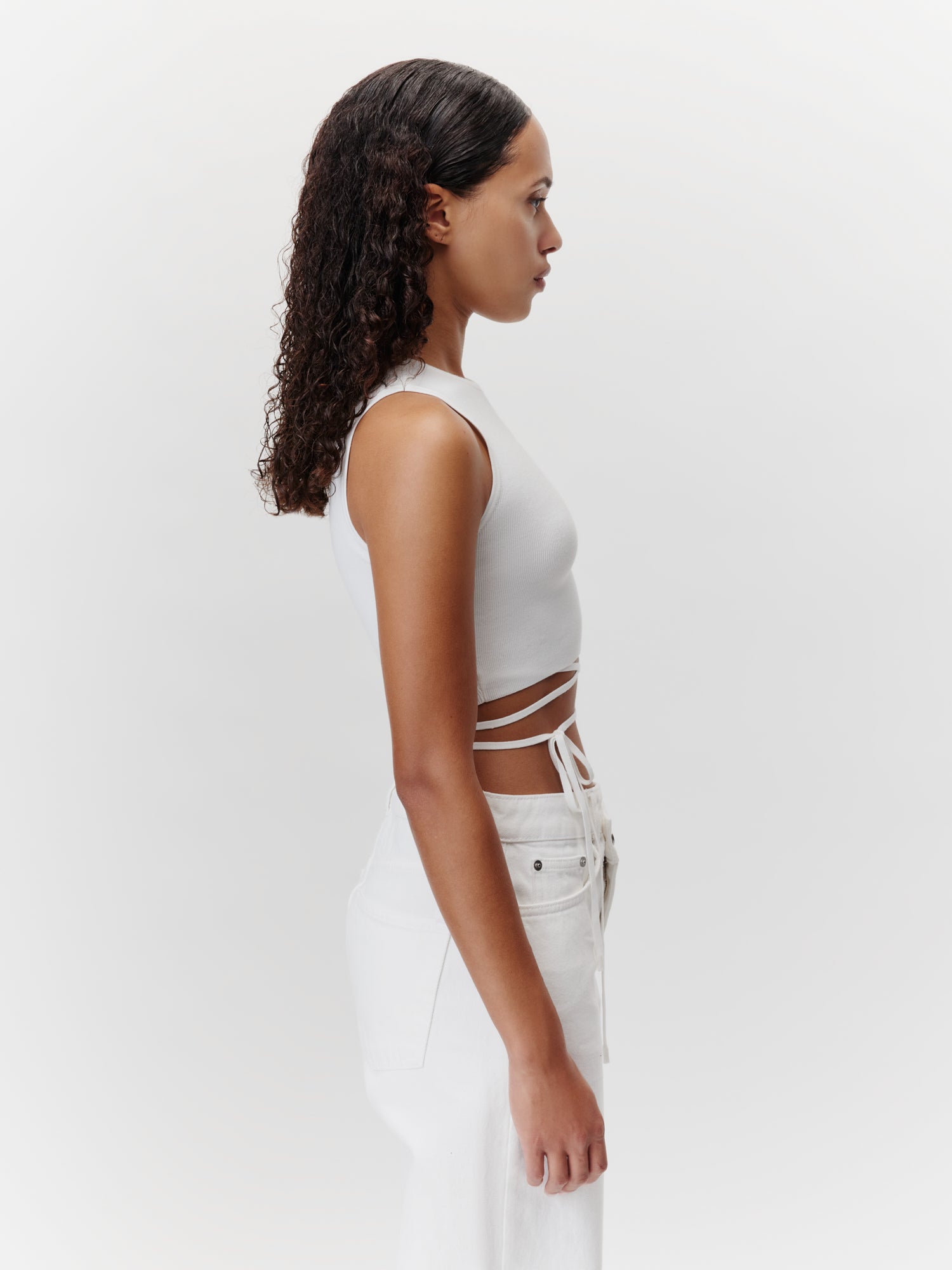Eine Frau mit lockigem Haar steht im Profil vor einem schlichten Hintergrund. Sie trägt das Leany Top von LeGer by Lena Gercke, ein ärmelloses weißes Crop-Top mit Wickelbändern, dazu hoch taillierte, figurbetonte weiße Hosen. Ihr Gesichtsausdruck ist neutral und ihre Haltung entspannt.
