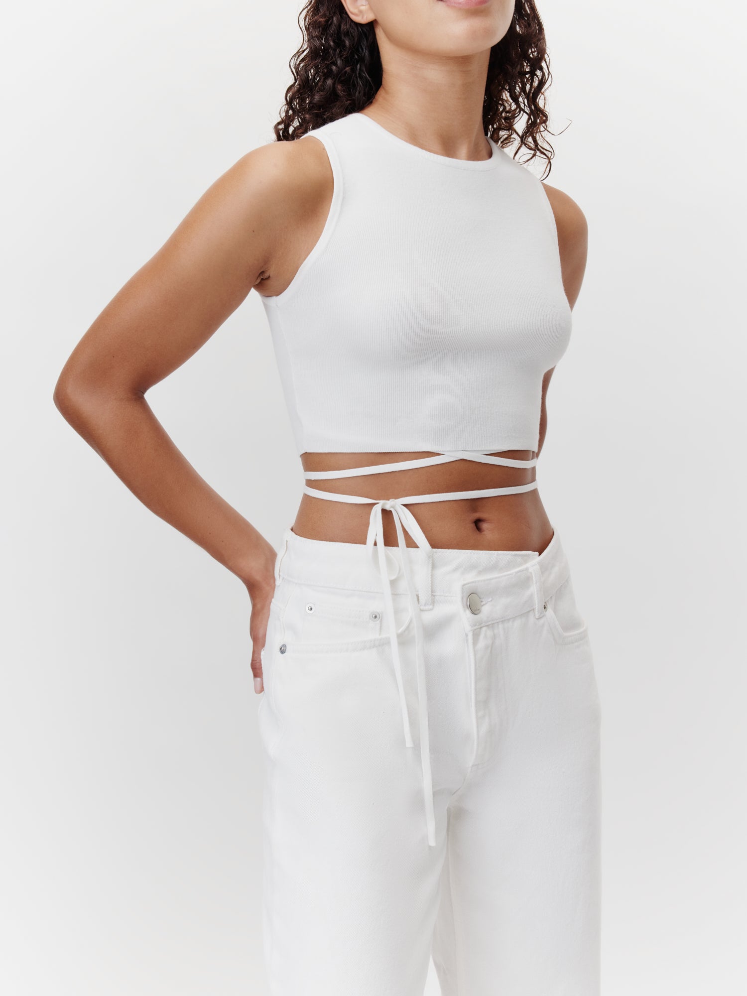 Eine Person trägt das Leany Top von LeGer by Lena Gercke, ein figurbetontes, ärmelloses weißes Crop-Top mit Wickelbändern, und kombiniert es mit weißen Jeans mit Taschen und Gürtelschlaufen. Die Person steht vor einem schlichten weißen Hintergrund und hat langes lockiges Haar.