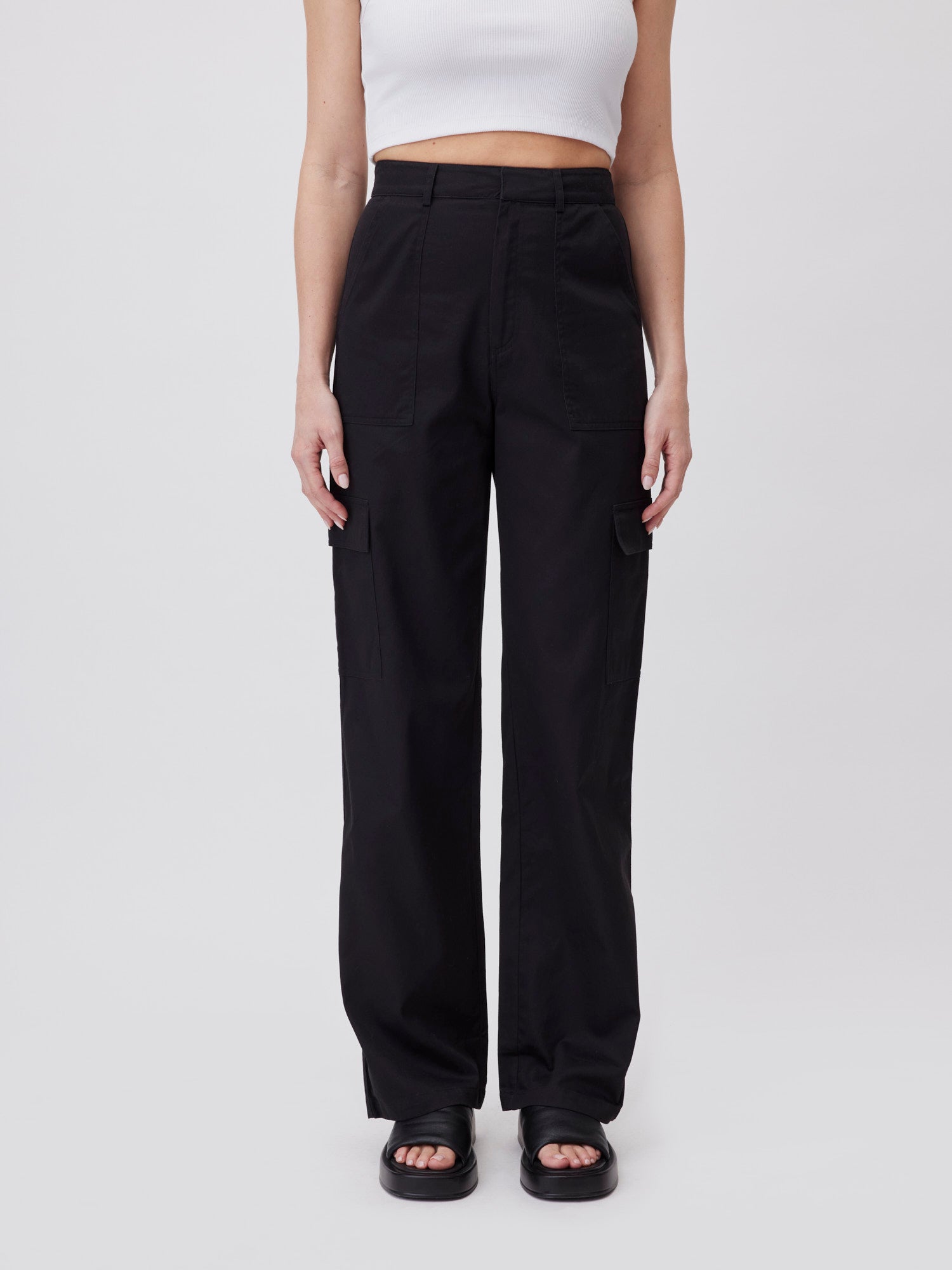 Eine Person trägt hoch taillierte schwarze Beysa-Hosen von LeGer by Lena Gercke in Lang/Maxi Länge, ein weißes kurzes Tanktop und schwarze Sandalen und steht vor einem schlichten weißen Hintergrund. Das Bild ist an den Schultern abgeschnitten.