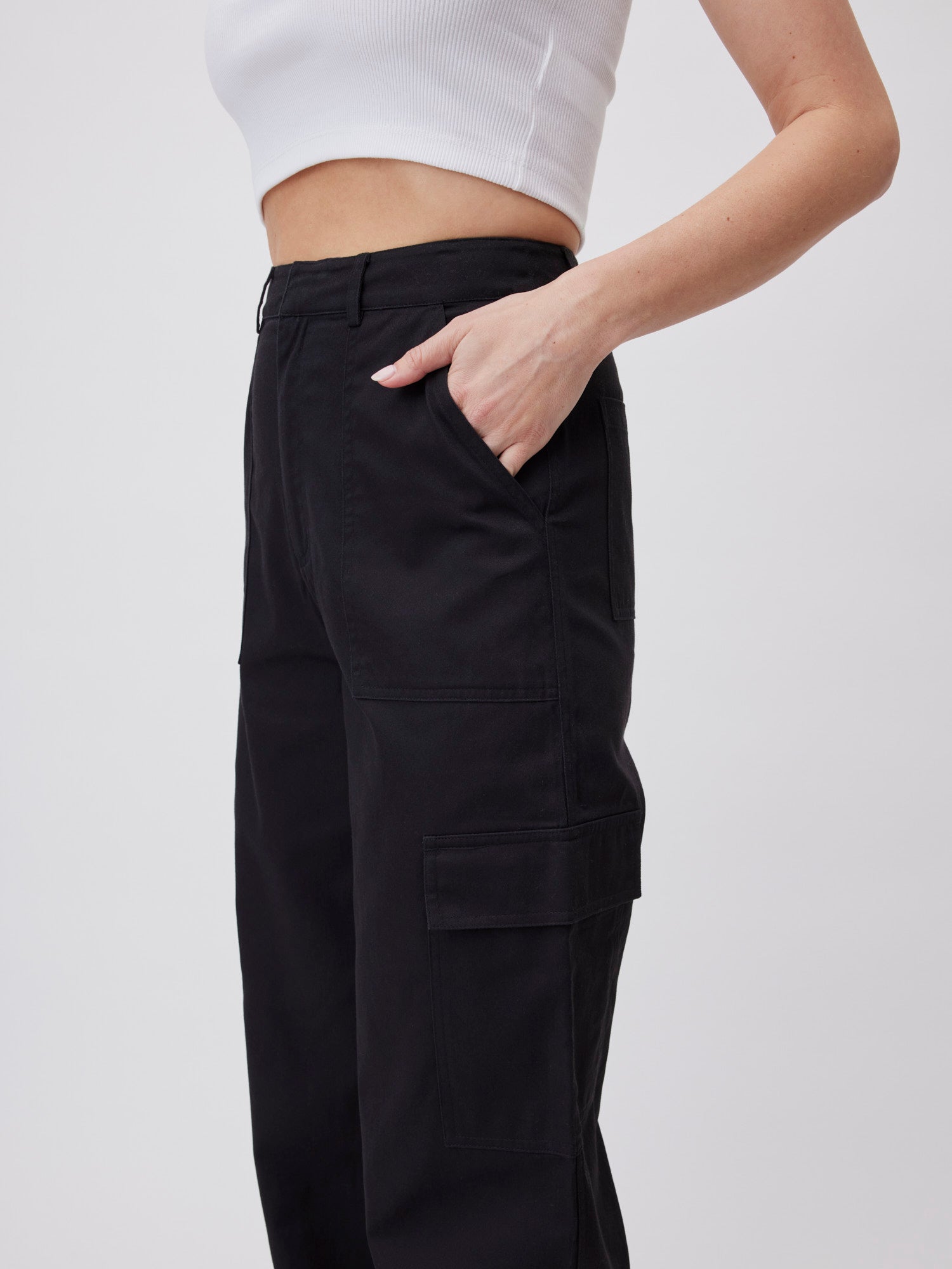 Eine Person präsentiert die LeGer by Lena Gercke Beysa Pants – eine hoch taillierte schwarze Cargohose in Lang/Maxi Länge – mit einem weißen gerippten Crop-Top und einer Hand in der Tasche. Der schlichte, helle Hintergrund unterstreicht die Besonderheiten des Outfits.